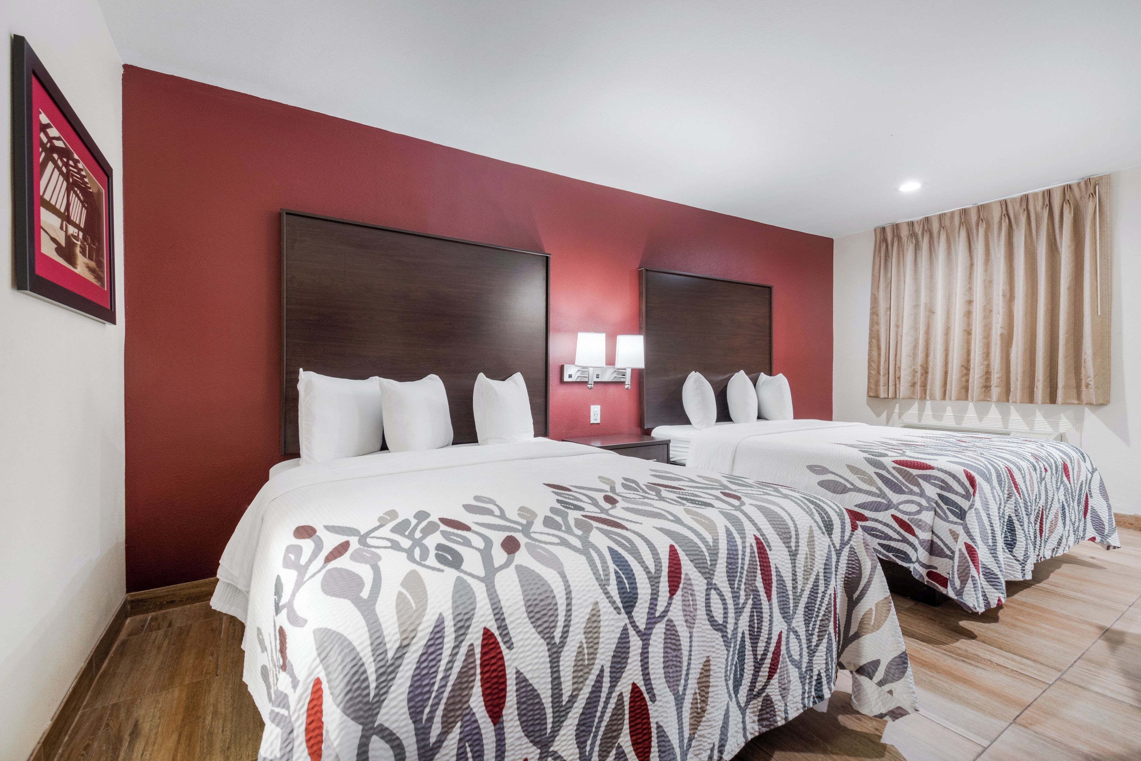 A hotel room with two beds, a red accent wall, and a large window with curtains.