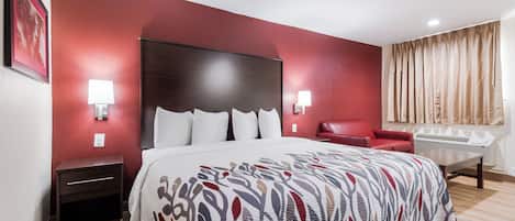 Quarto de hotel com uma cama grande, uma cadeira vermelha, uma escrivaninha e uma janela com cortinas.