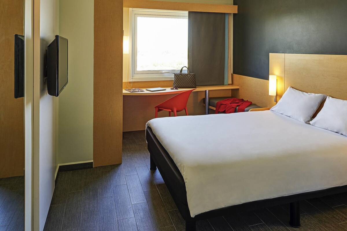 A hotel room with a bed, a desk with a chair, a television, and a window.