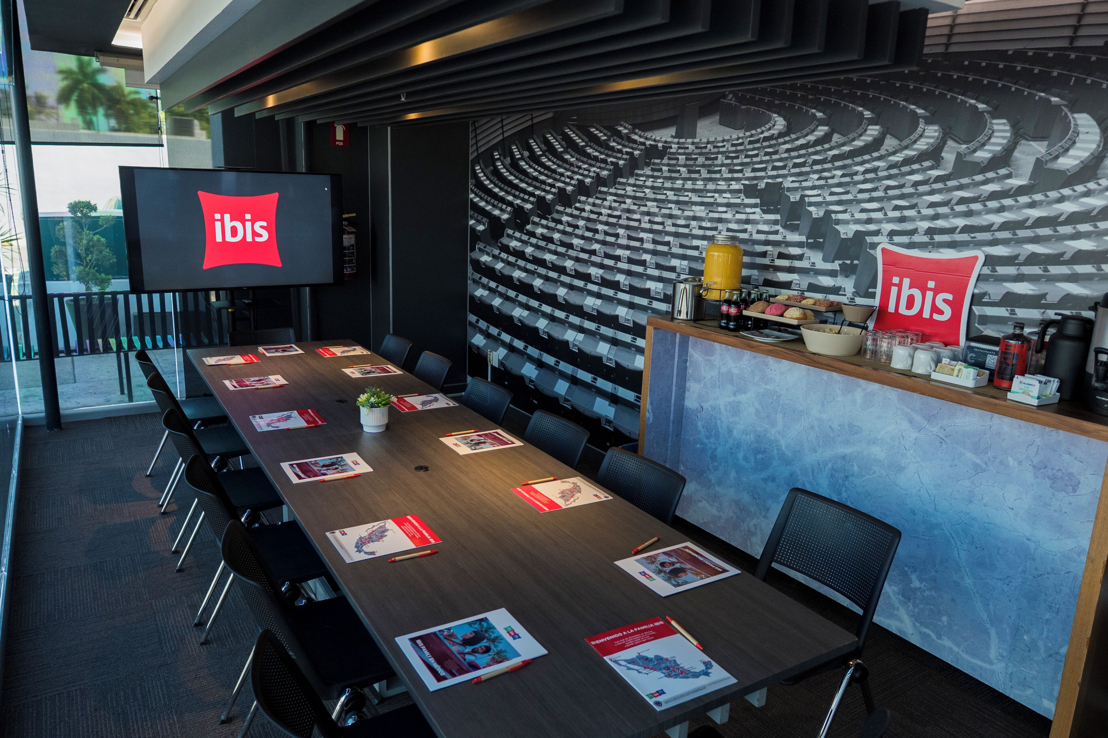 A conference room with a long table, chairs, a screen displaying the ibis logo, and a wall mural of an auditorium.