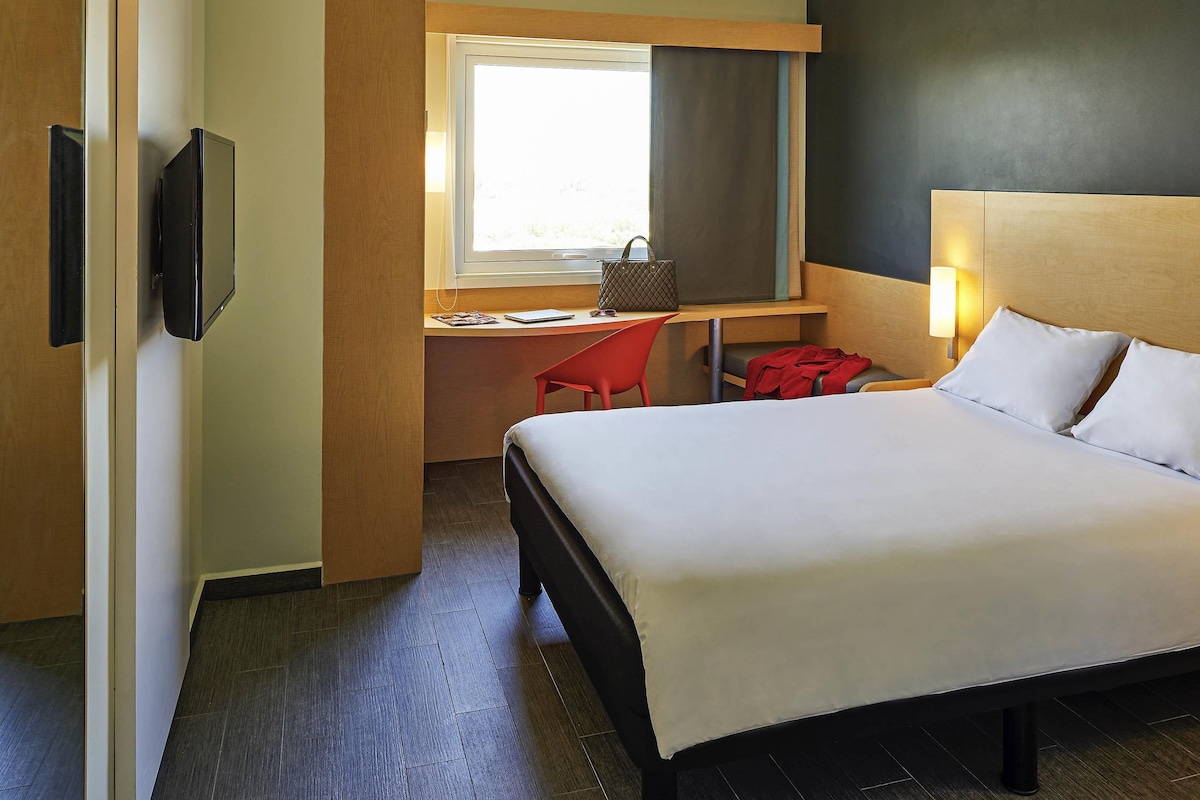 Une chambre d’hôtel avec un lit, un bureau et une chaise, une télévision et une fenêtre.