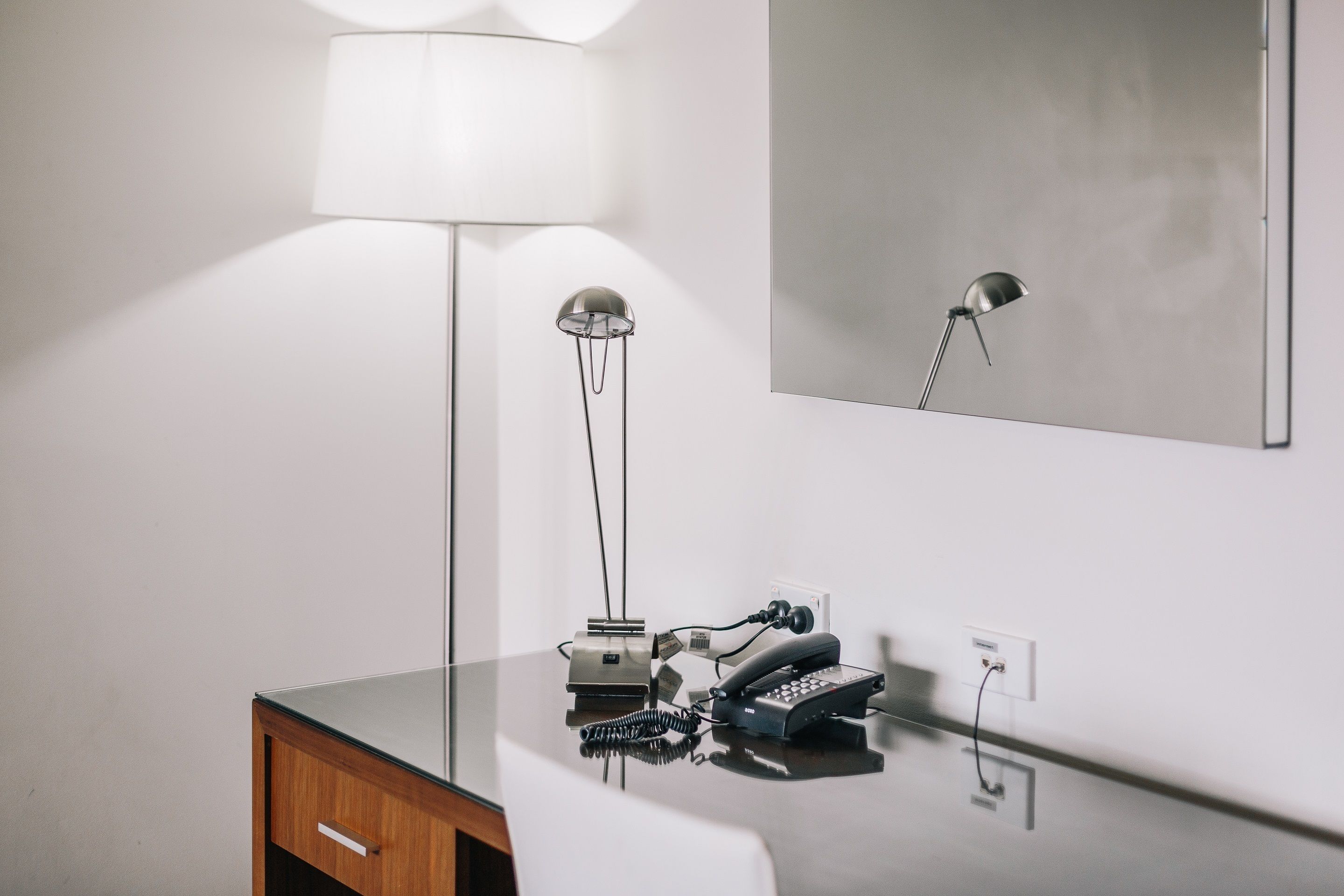 A modern hotel room with a glass-top desk, a lamp, a telephone, and a mirror on the wall.