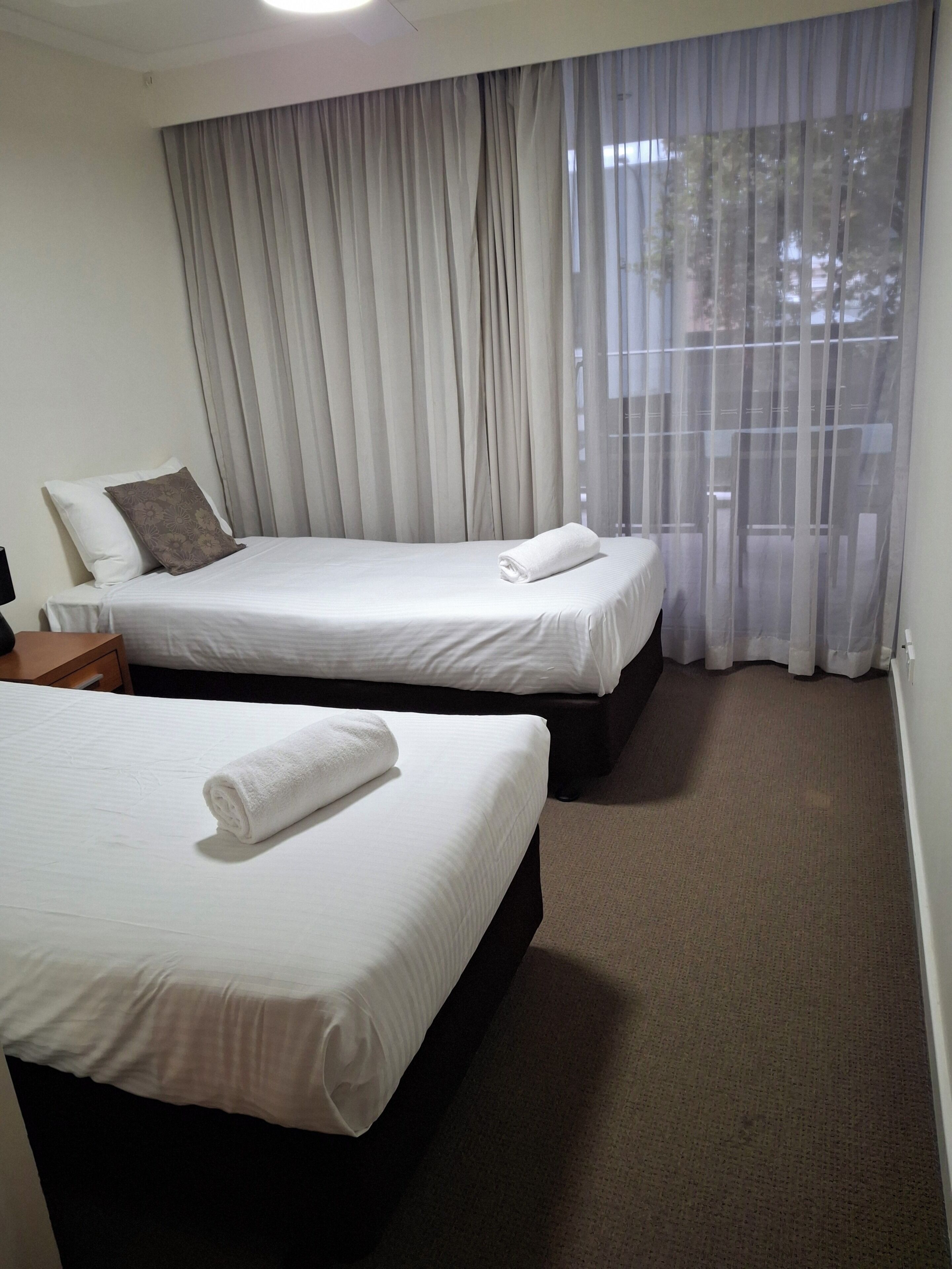 A hotel room with two beds, a window with sheer curtains, and a carpeted floor.