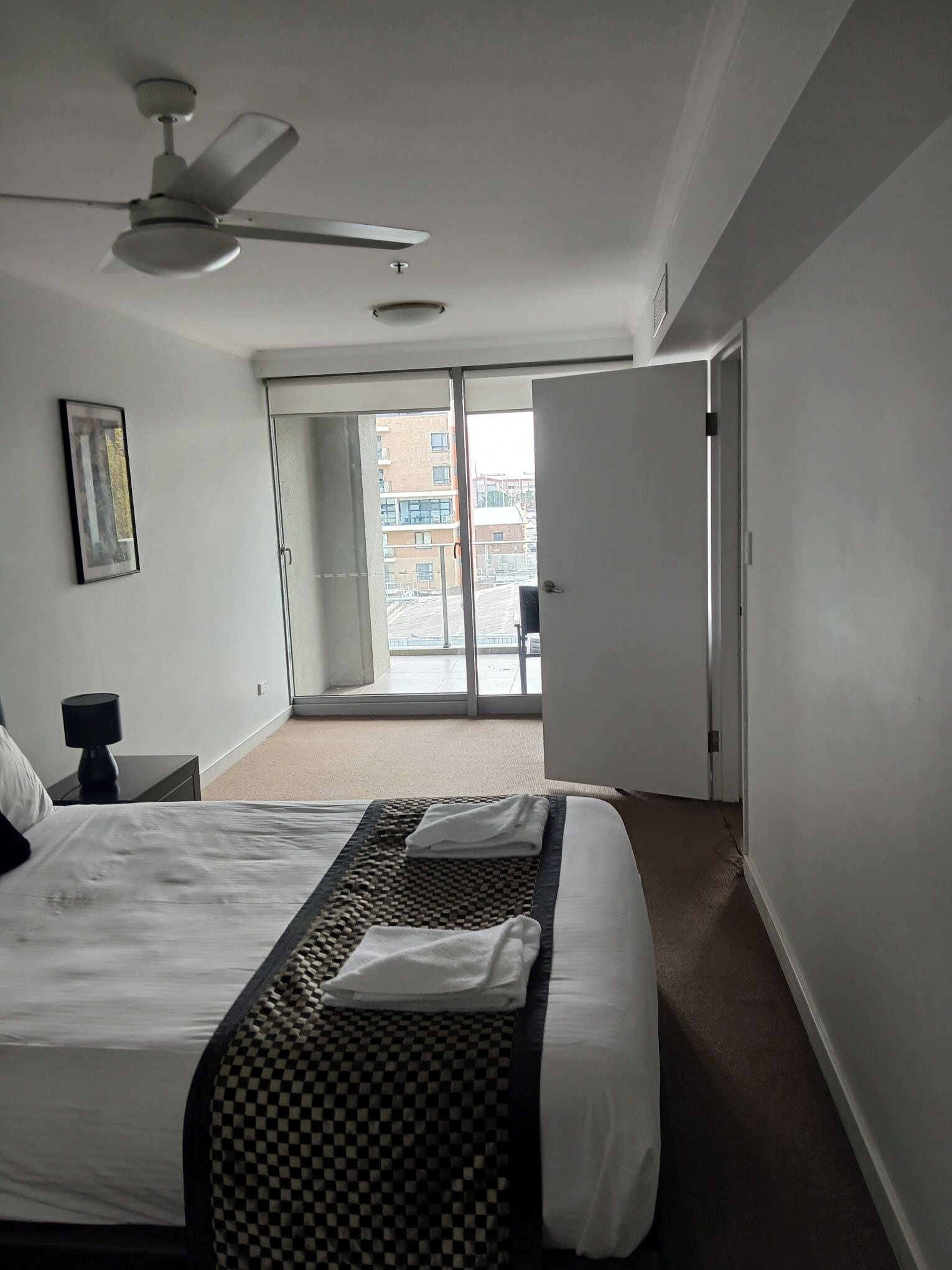 Hotellihuone, jossa on sänky, kattotuuletin, ovi, joka johtaa parvekkeelle, ja seinällä oleva kehystetty kuva.