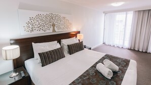 Una camera d'albergo con un letto grande, un comodino con una lampada, un quadro appeso al muro e una finestra con tende.