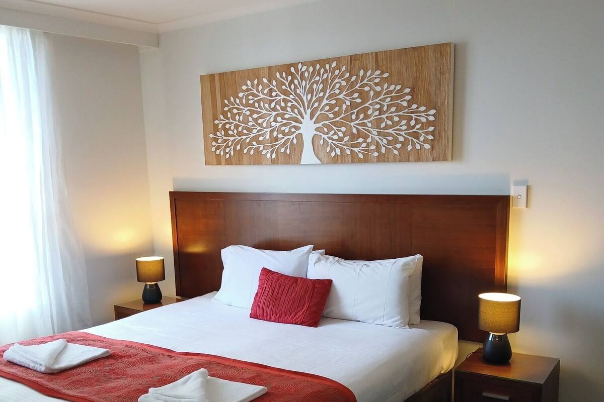 Ein Hotelzimmer mit einem großen Bett, zwei Nachttischen mit Lampen, einem Holz-Kopfteil, einer Wanddekoration mit Baummotiv und einer roten Tagesdecke.