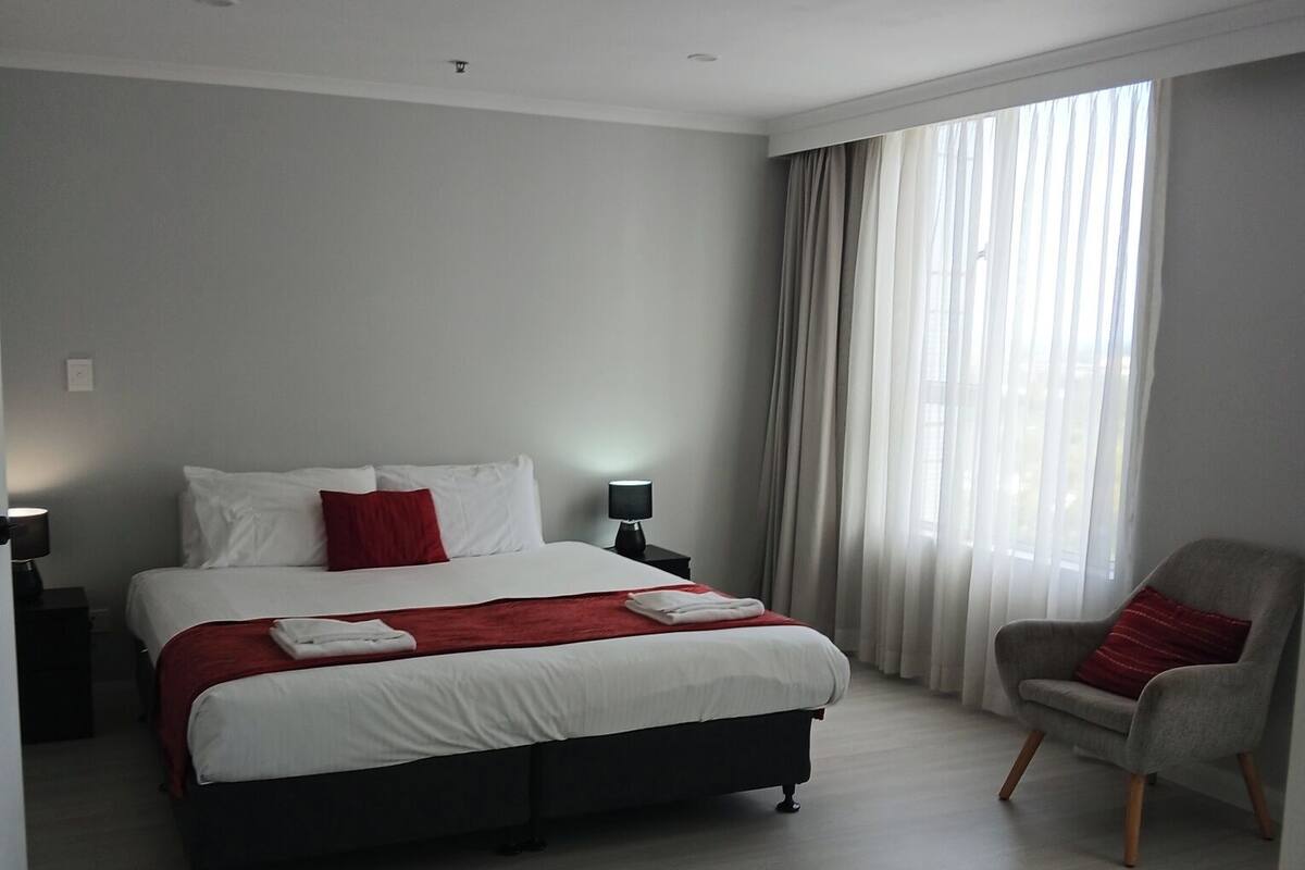 Ein modernes Hotelzimmer mit einem großen Bett, einem Sessel, einem Nachttisch und einem Fenster mit Vorhängen.