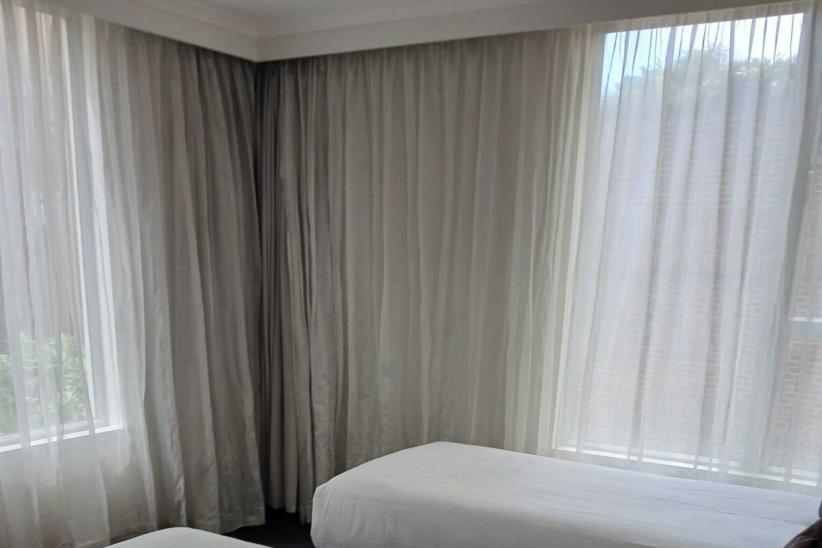 Ein Hotelzimmer mit zwei Betten, einem großen Fenster mit durchsichtigen Vorhängen und Blick ins Grüne.