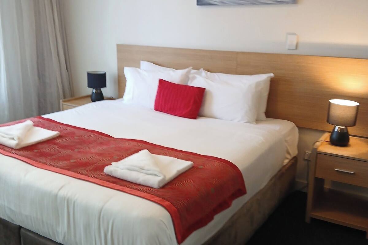 Ein Hotelzimmer mit Bett, Nachttisch, Lampe, Gemälde und einem fensterbepflanzten Fenster.