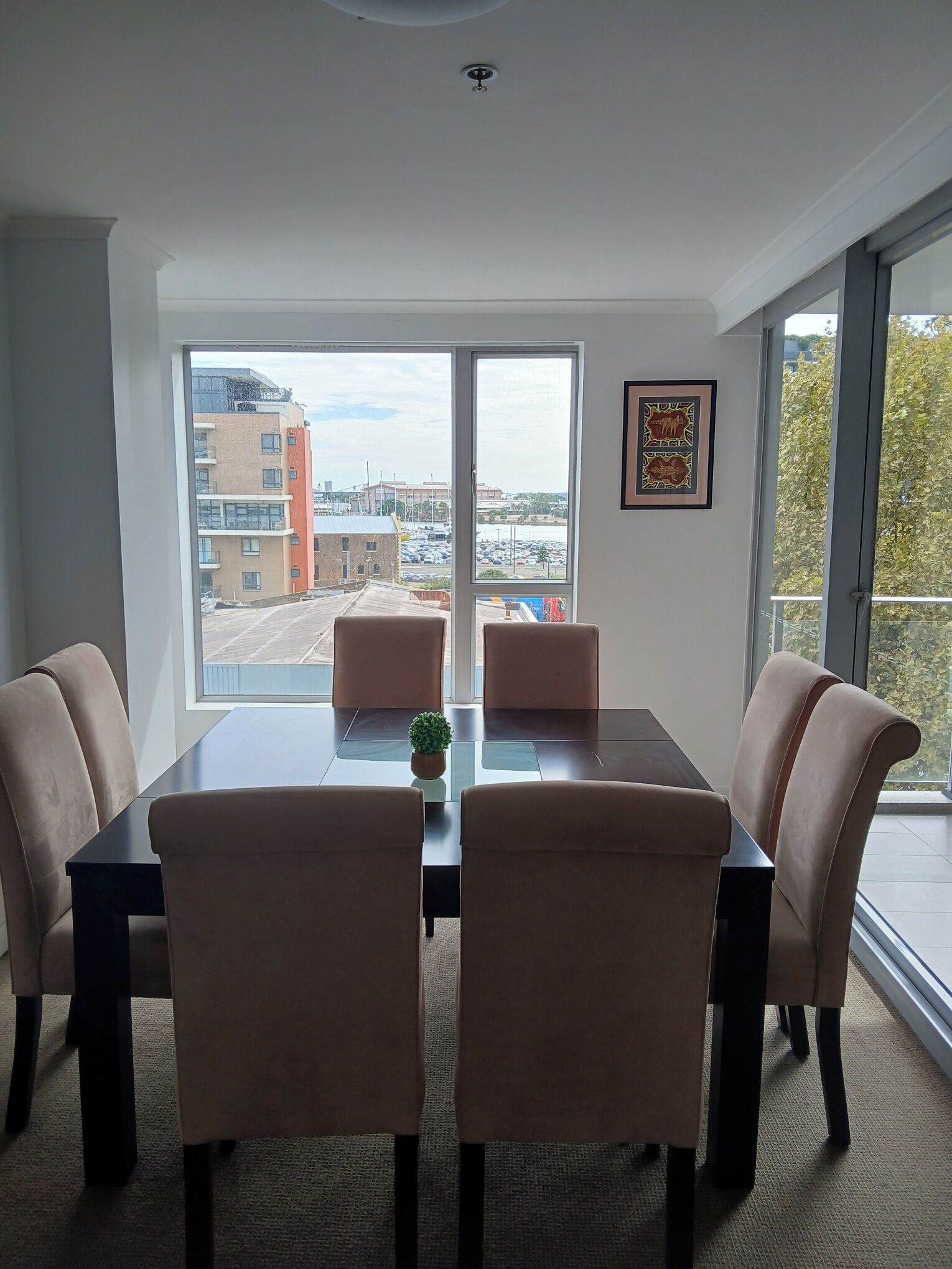 Uma sala de jantar moderna com uma mesa de vidro, quatro cadeiras estofadas e vista para edifícios e um parque de estacionamento através de grandes janelas.