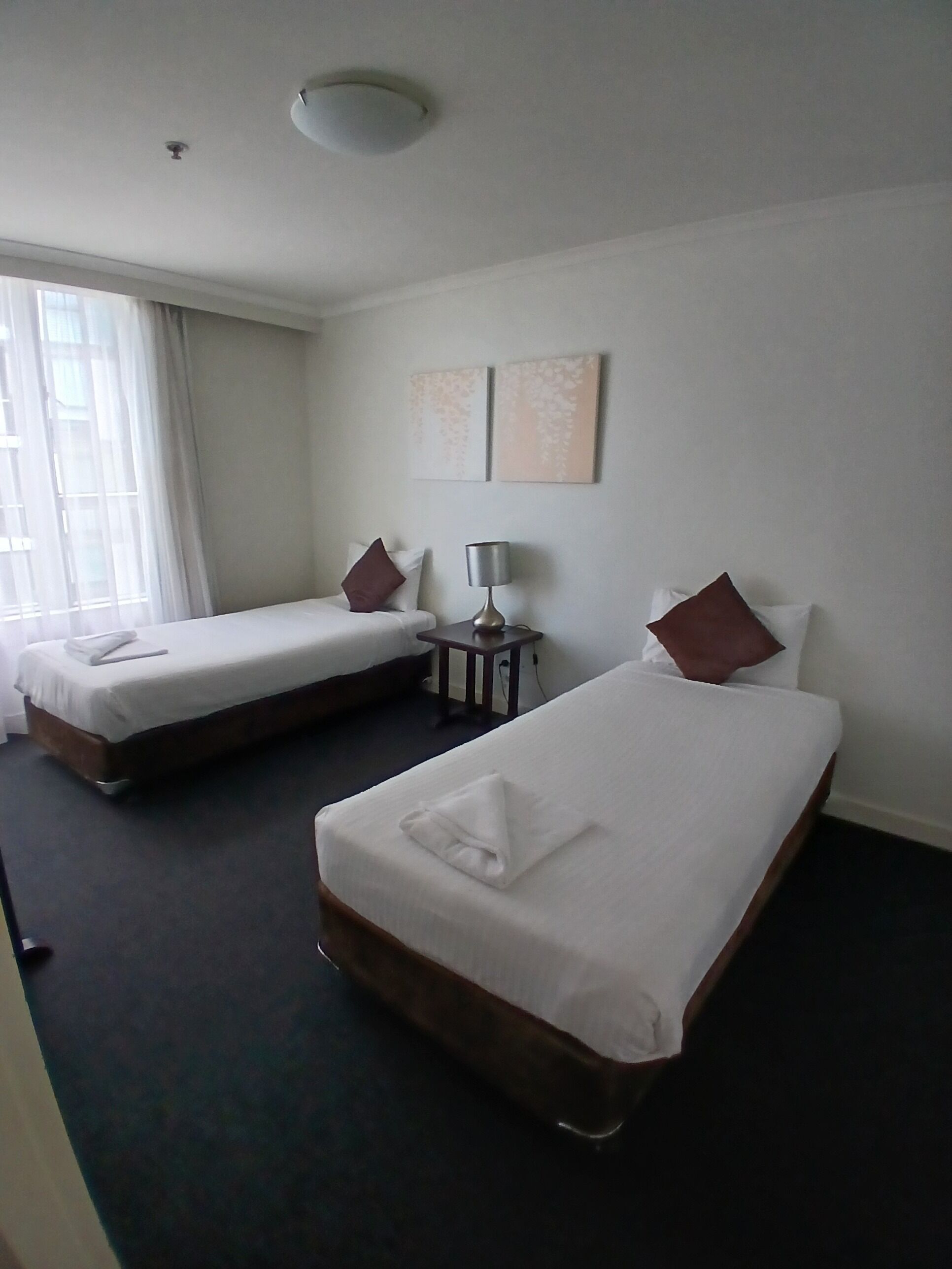 Una habitación de hotel con dos camas individuales, una mesita de noche con lámpara y una ventana con cortinas.