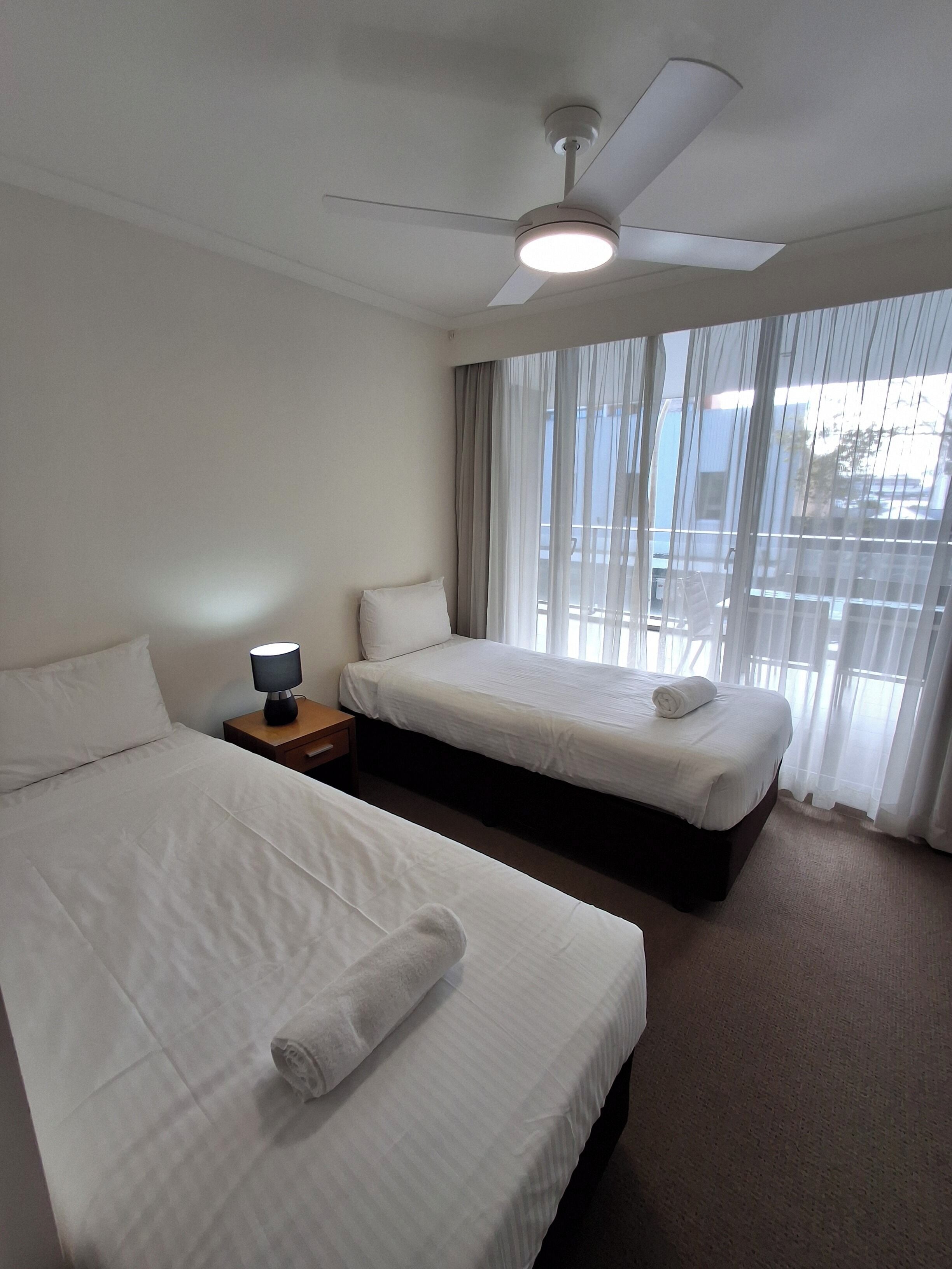 A hotel room with two beds, a ceiling fan, a nightstand with a lamp, and a view through sheer curtains.