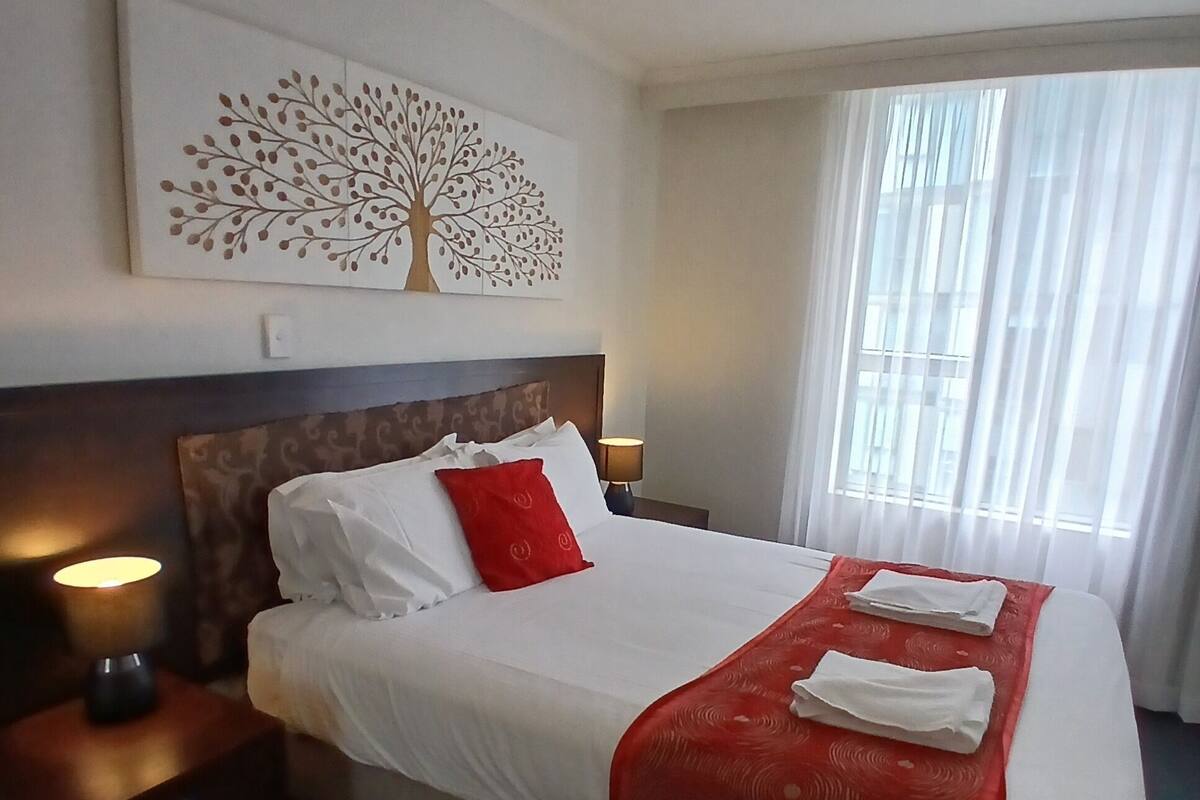 Ein Hotelzimmer mit Bett, Nachttisch mit Lampe, einem Fenster mit Vorhängen und einem Wandbild eines Baumes.