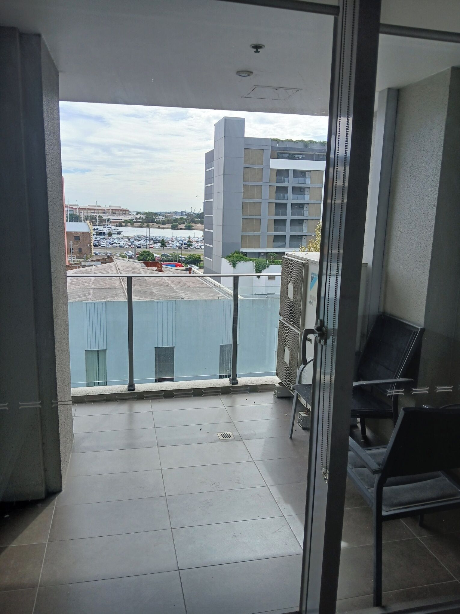 Een balkon met uitzicht op een parkeerplaats en gebouwen, een stoel en een ventilator.