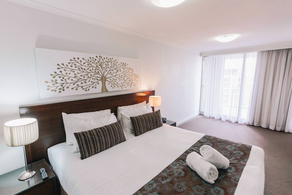 Ein Hotelzimmer mit einem großen Bett, einem Nachttisch mit Lampe, einem Wandbild und einem Fenster mit Vorhängen.