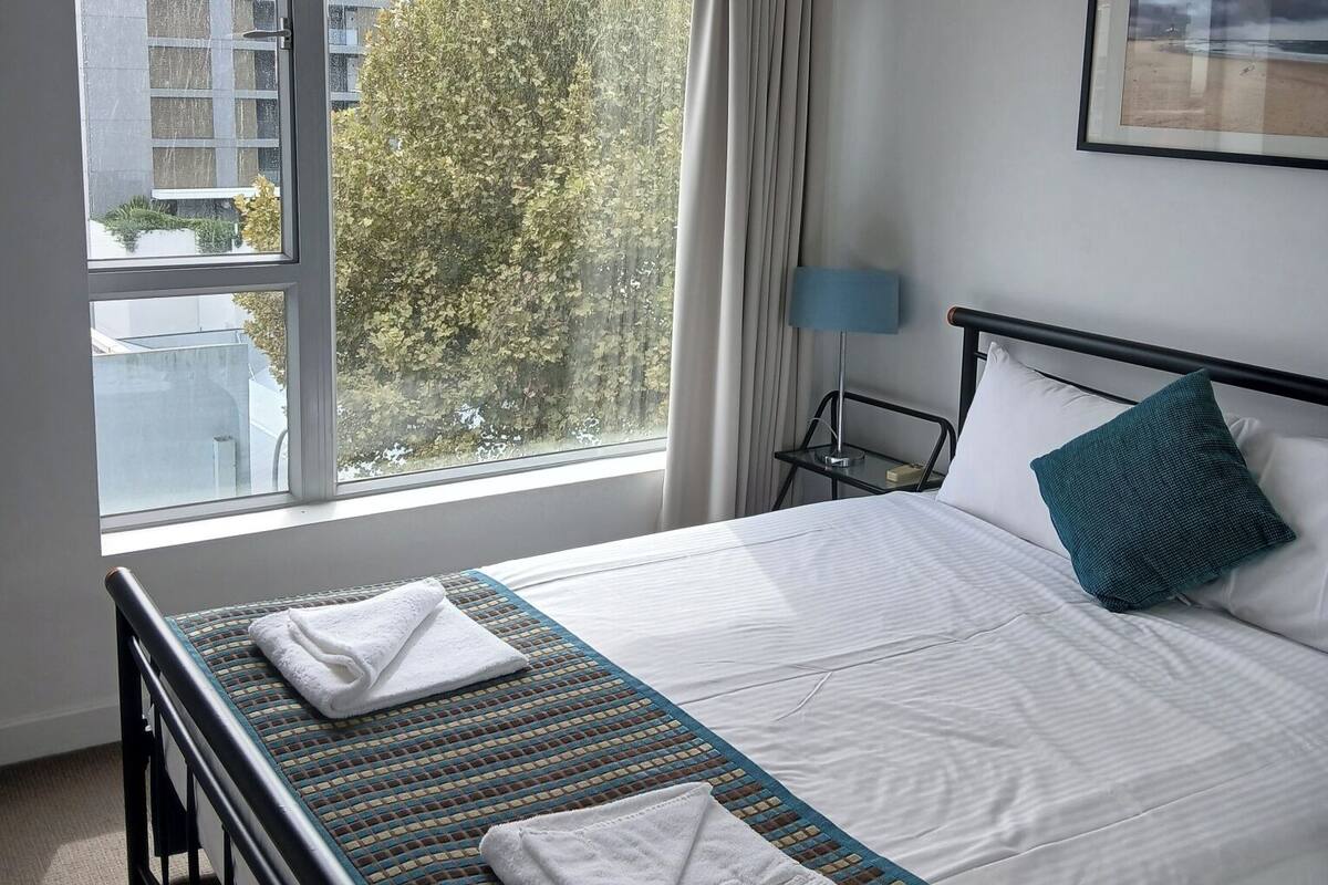 Ein Hotelzimmer mit Bett, Deckenventilator, Fenster mit Blick auf Gebäude und Bäume, Nachttisch mit Lampe sowie ein gerahmtes Bild an der Wand.