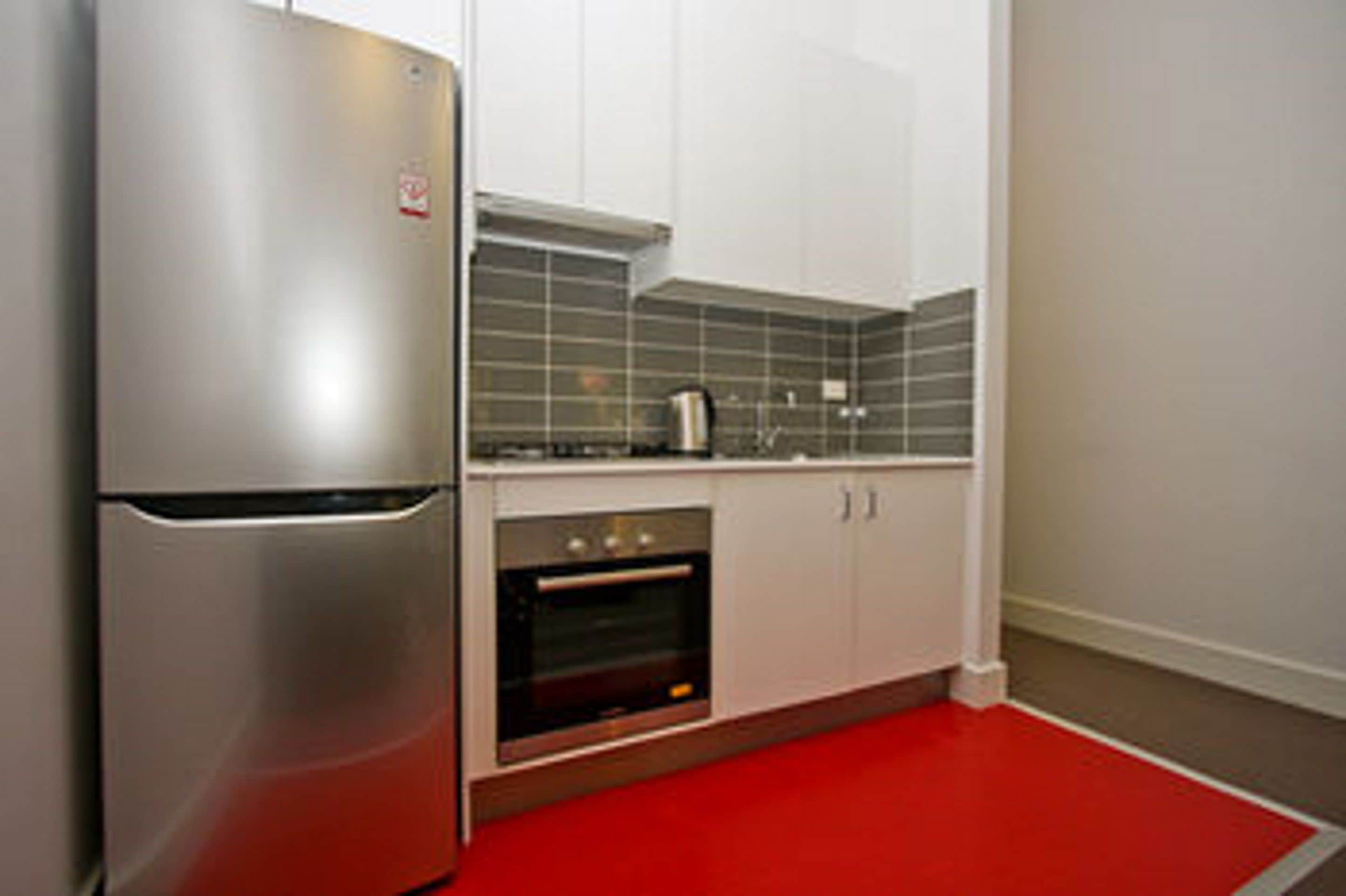 Una cocina con refrigerador de acero inoxidable, horno empotrado, gabinetes blancos y un tapete rojo.