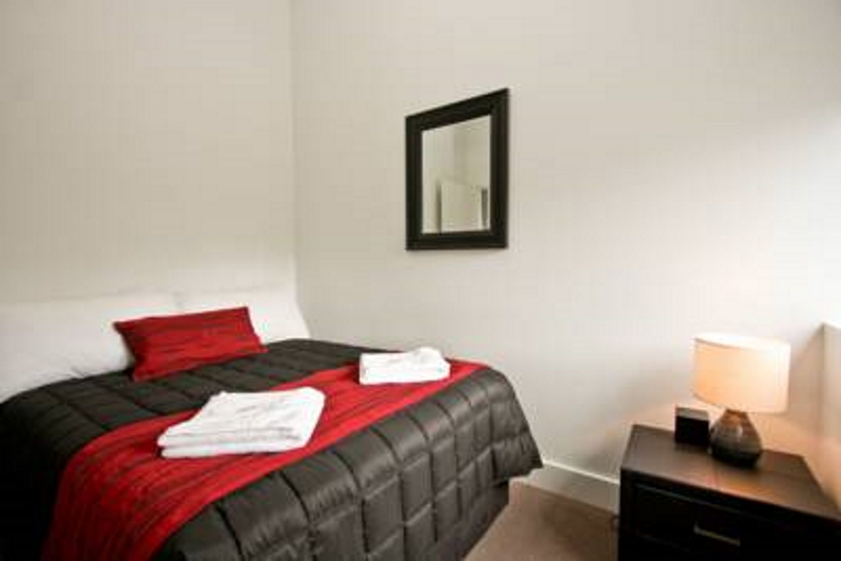 A neatly made bed with a black and red bedspread, a white pillow, and a red accent pillow. There is a bedside table with a lamp and a small decorative item. A mirror is mounted on the wall above the bed.
