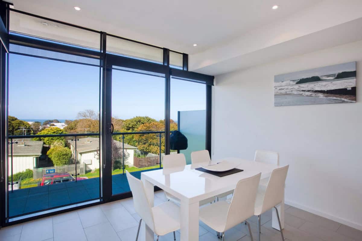 A modern dining room with a large table and chairs, large windows with a view of the sea and surrounding landscape.