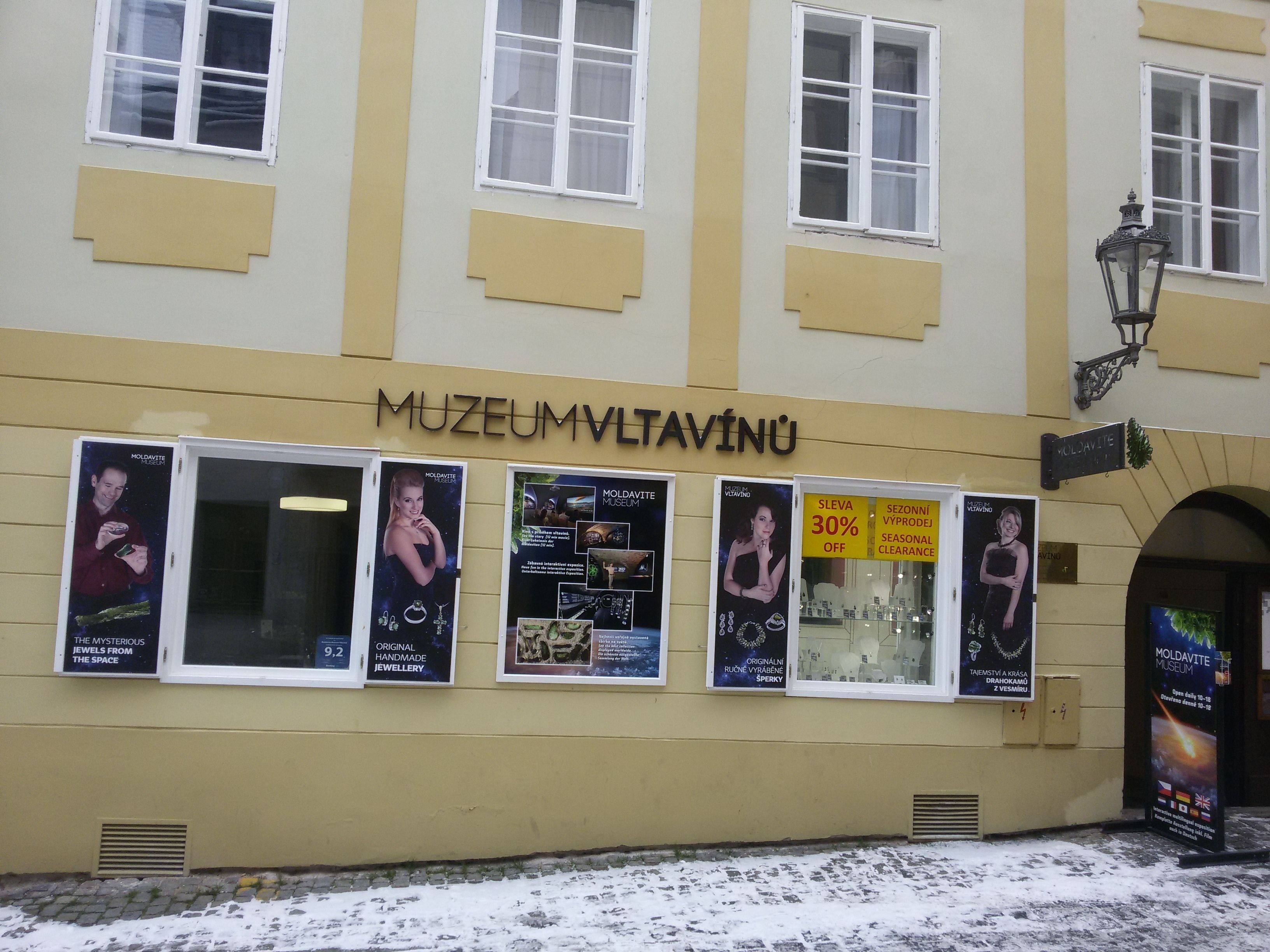A building with the sign "MUZEUMMVLTAVINU" indicating a museum. There are multiple posters advertising the museum's exhibits and a seasonal clearance sale.