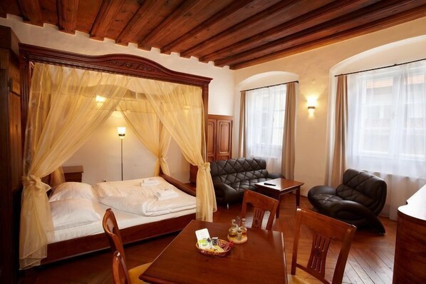 A hotel room with a large bed, a sitting area with a leather sofa, a dining table, and a small table with a basket of snacks.