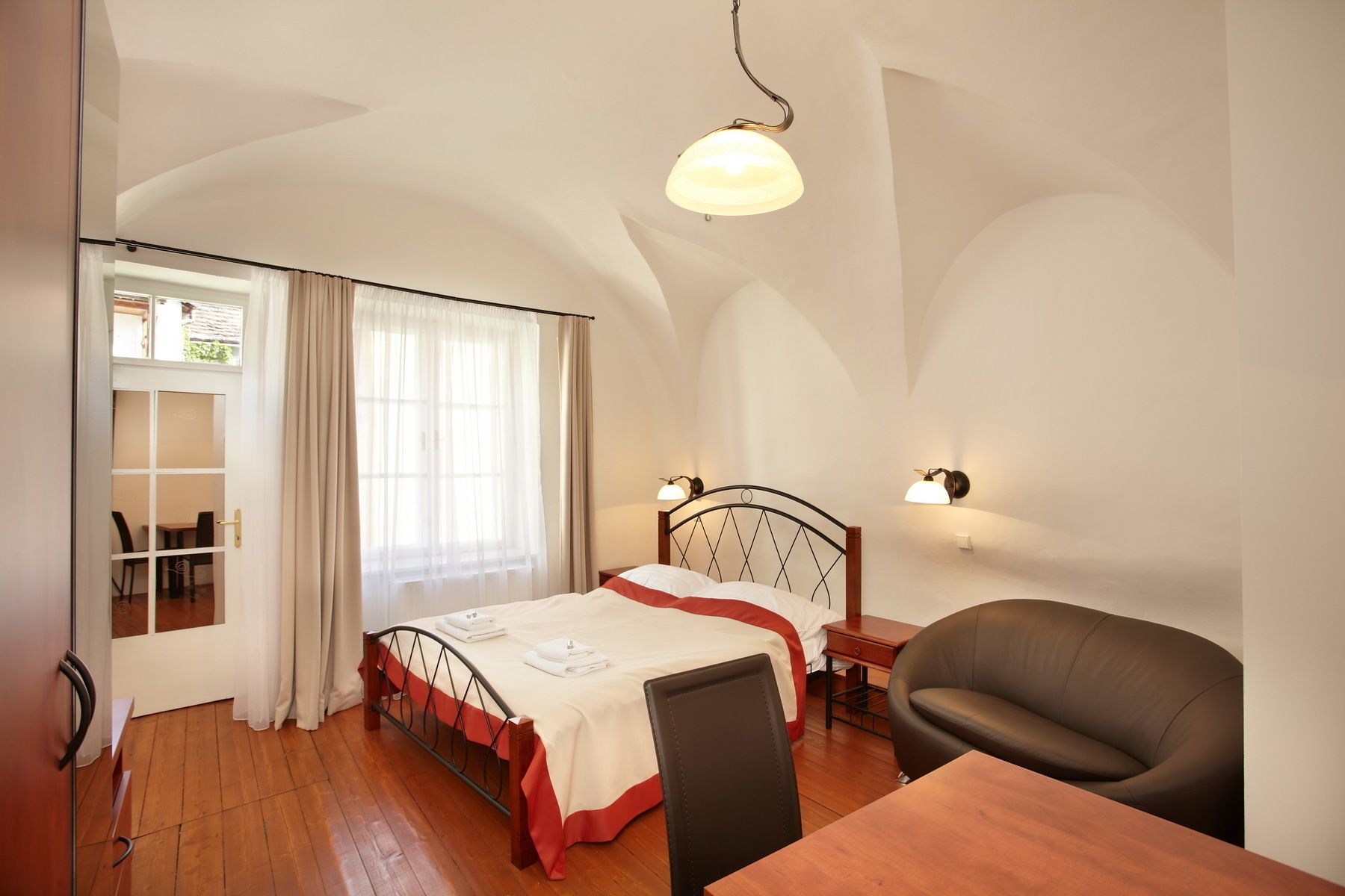 A bedroom with a double bed, a wooden desk, a chair, and a window with curtains.