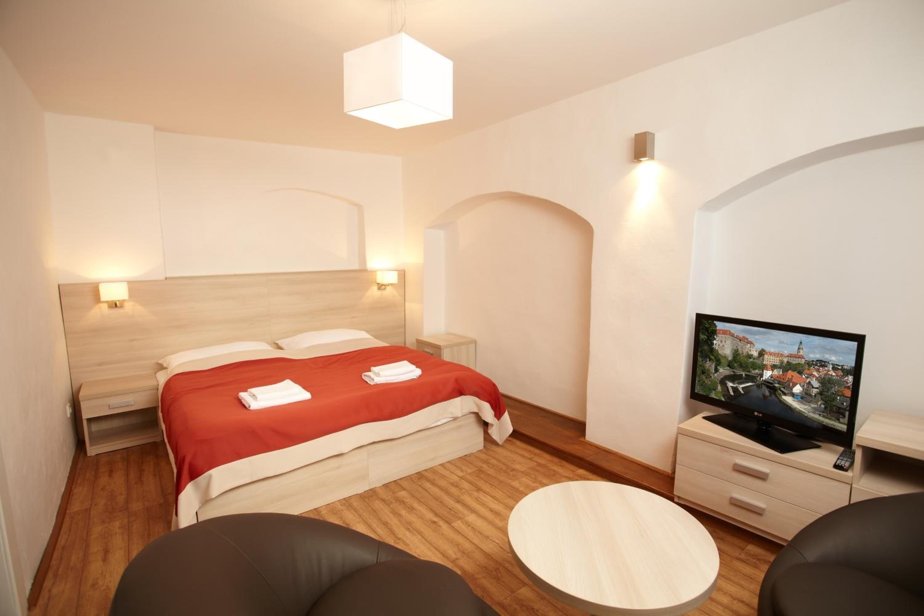 A modern hotel room with a wooden floor, a bed with a red blanket, a TV on a wooden cabinet, and a round table.