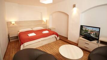 A modern hotel room with a wooden floor, a bed with a red blanket, a TV on a wooden cabinet, and a round table.