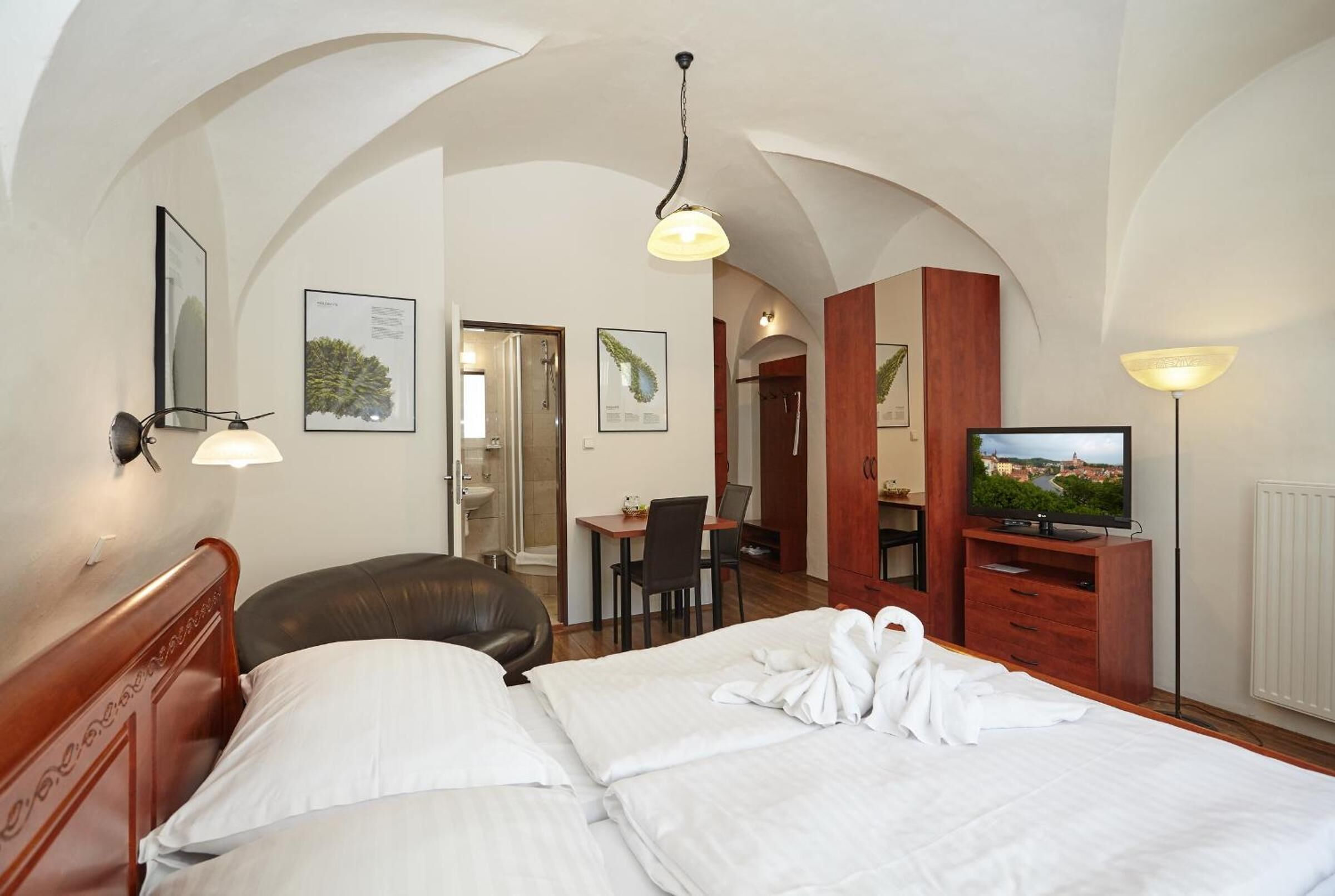A hotel room with a bed, a desk, a chair, a television, and a bathroom visible through an arched doorway.