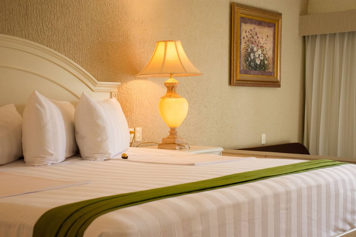 A neatly made bed with white bedding and pillows, a bedside lamp, a framed floral painting on the wall, and a green throw blanket.