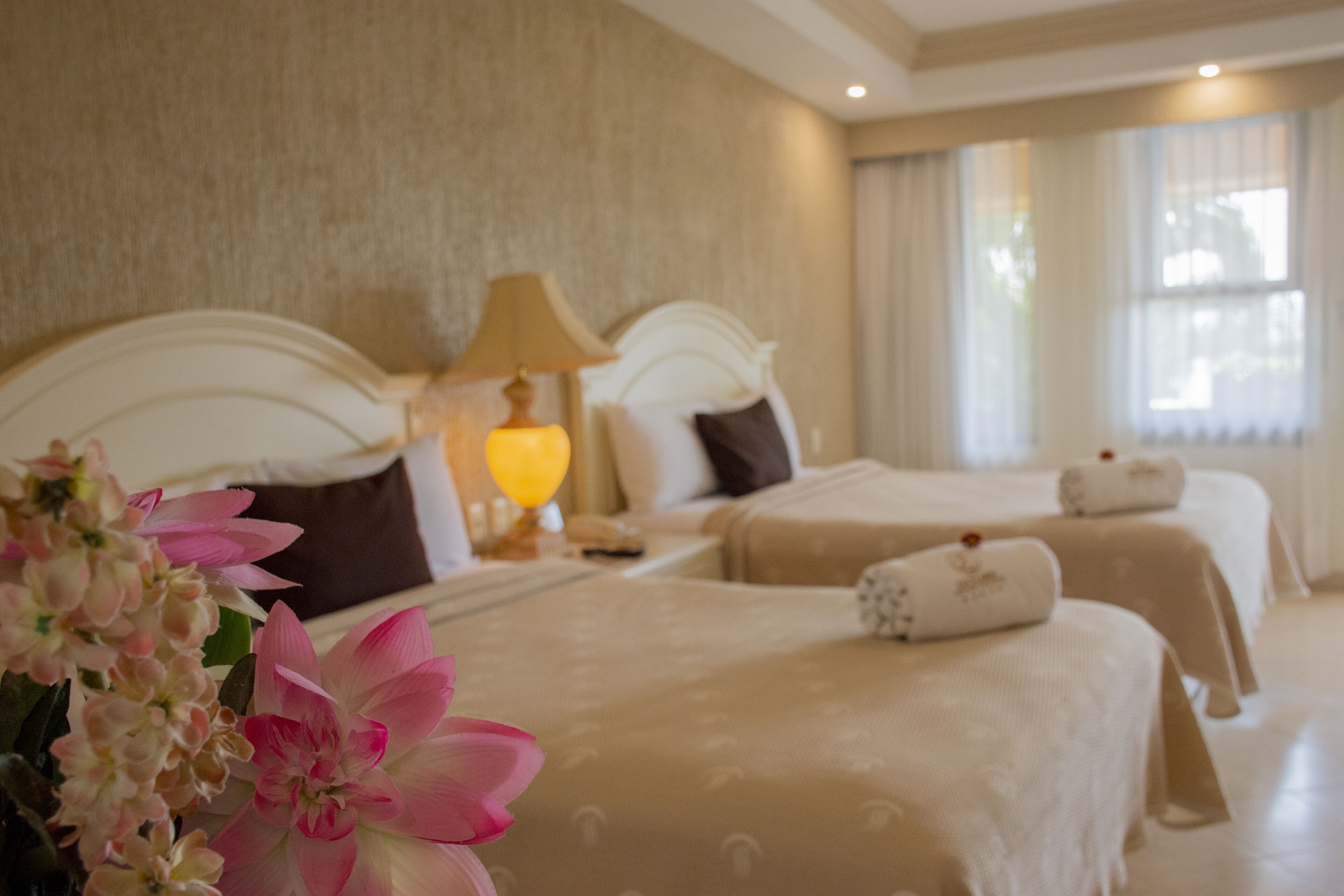 A hotel room with two beds, a nightstand with a lamp, and a large window with curtains.