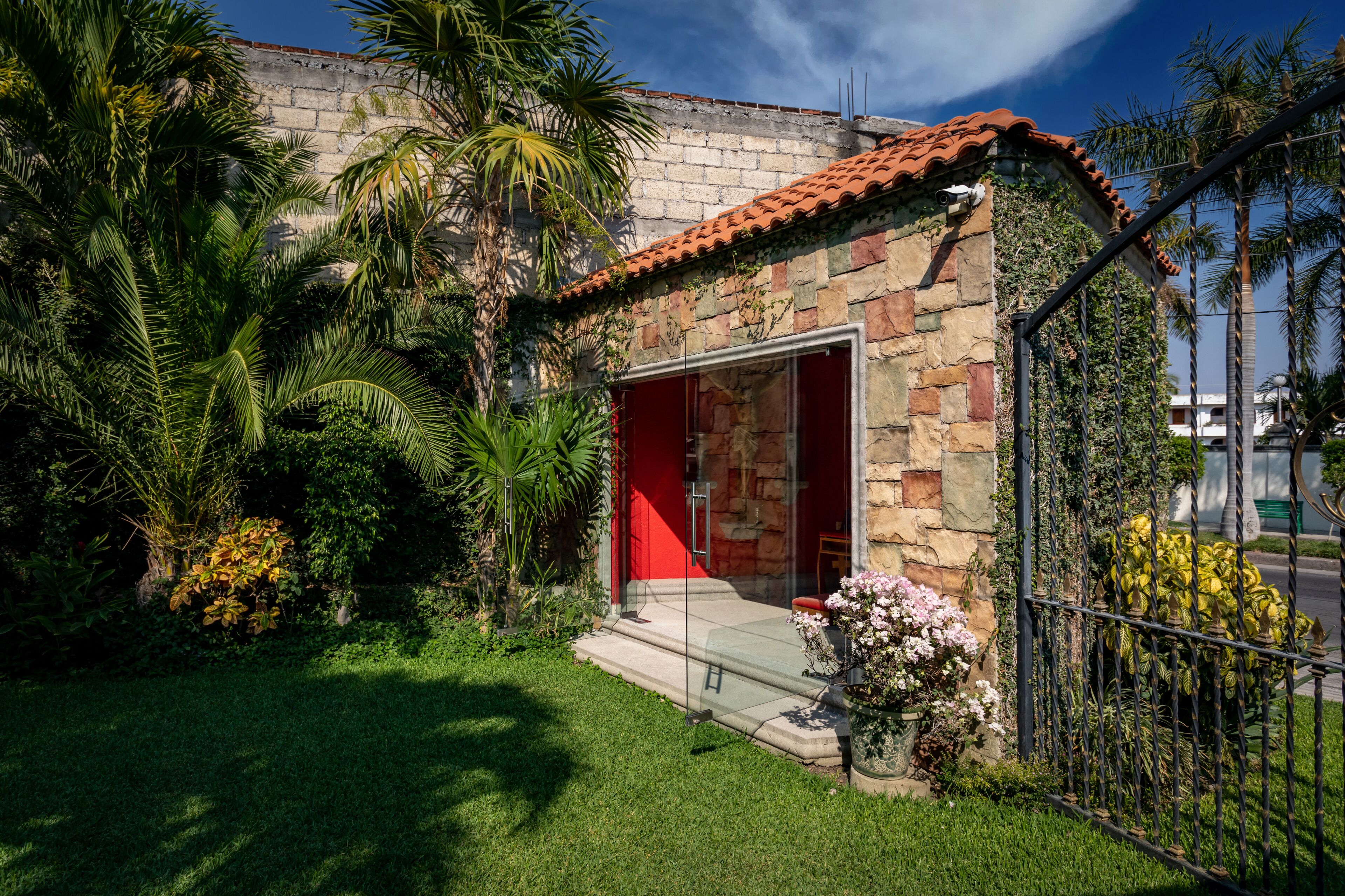 Un pequeño edificio de piedra con una puerta roja, rodeado de abundante follaje y palmeras.
