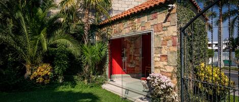 Un pequeño edificio de piedra con una puerta roja, rodeado de abundante follaje y palmeras.