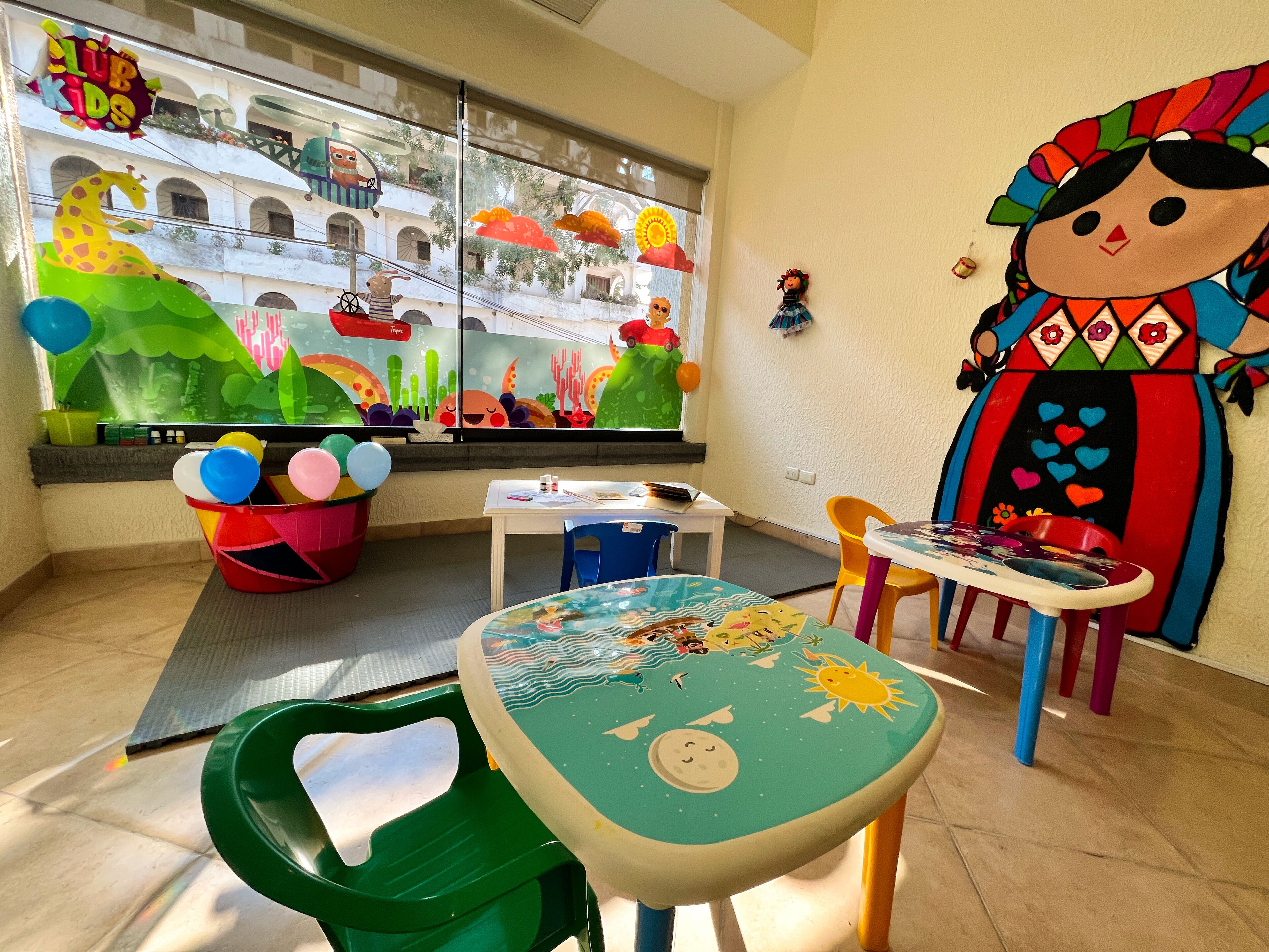 A children's playroom with colorful decorations, a large mural, and various play tables and chairs.