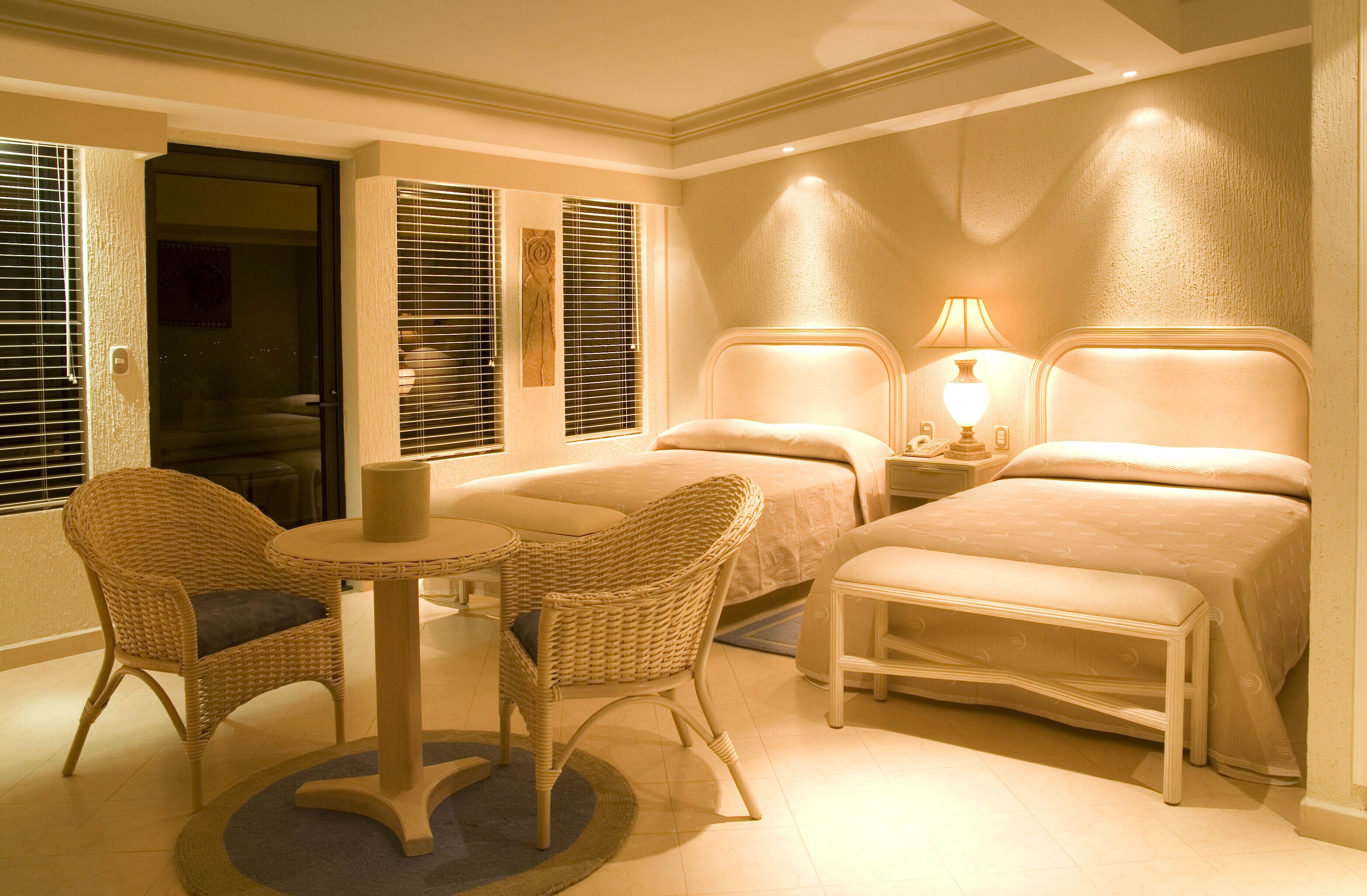 Habitación de hotel con dos camas, una mesita, dos sillas de mimbre y una mesita de noche con lámpara.