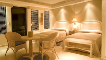 Habitación de hotel con dos camas, una mesita, dos sillas de mimbre y una mesita de noche con lámpara.