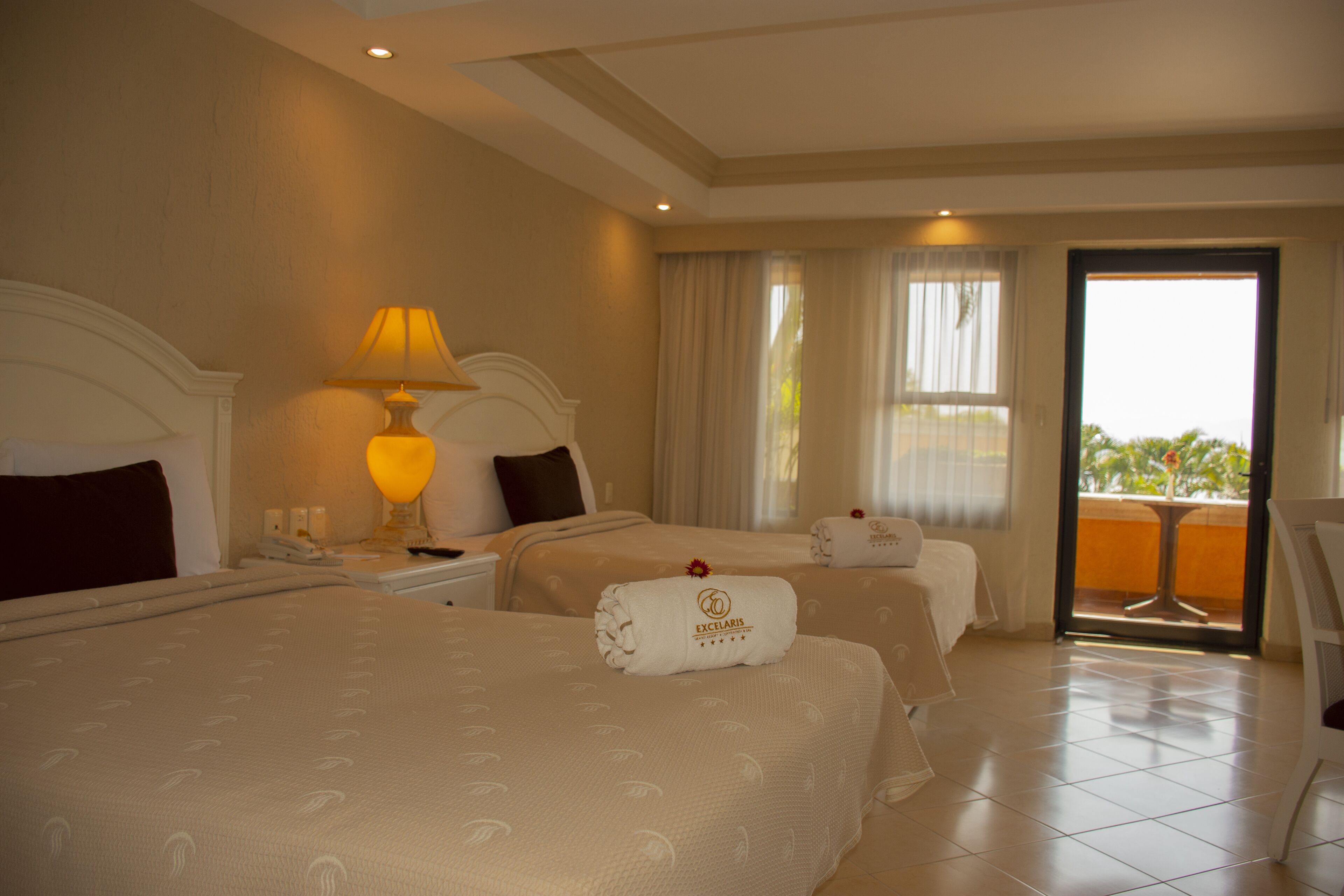 Habitación de hotel con dos camas, una mesita de noche con lámpara, una ventana con cortinas y vistas al exterior.