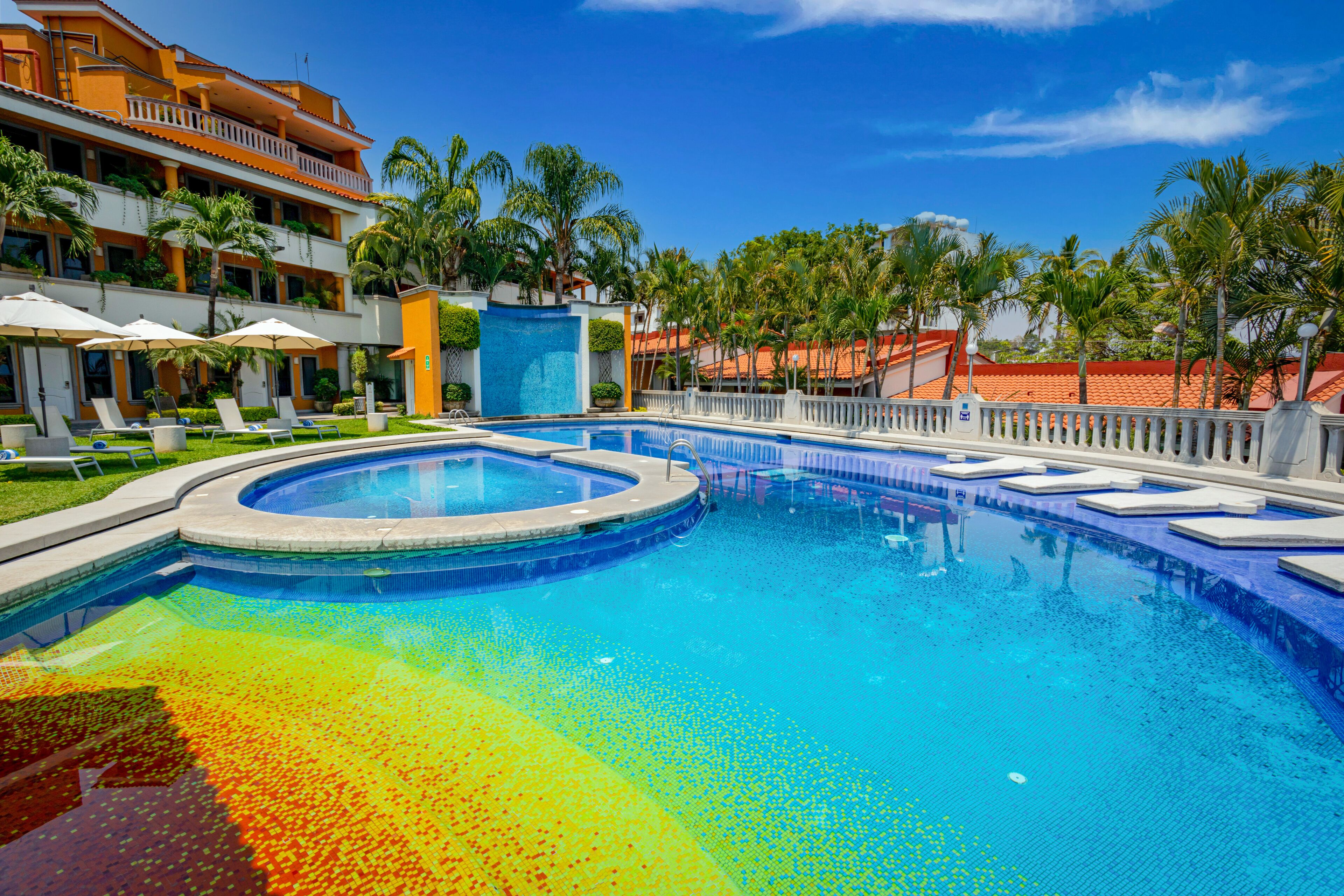 Una piscina de diseño circular, rodeada de sillas de descanso y sombrillas, con un edificio de balcones y palmeras al fondo.