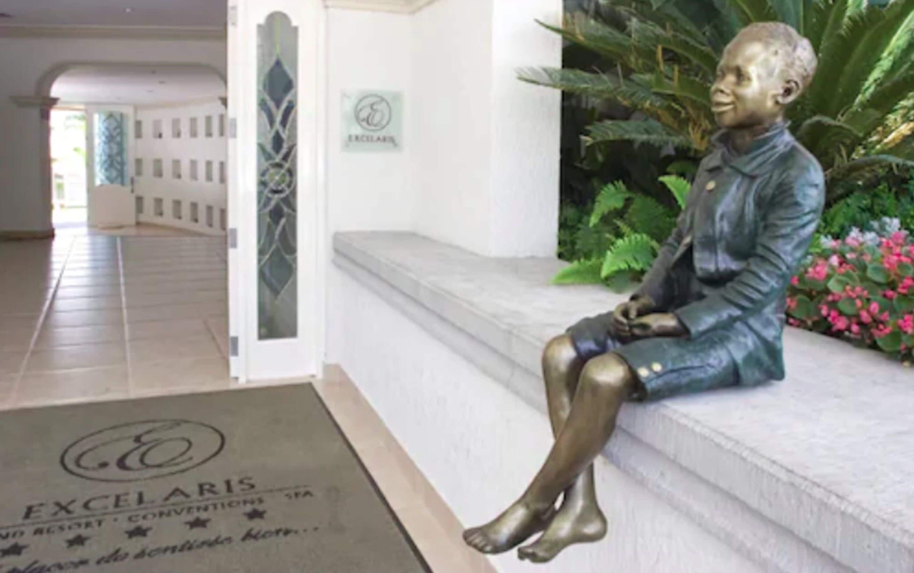 A statue of a seated figure on steps, a sign reading "EXCELARIS," and a doormat with the same name.