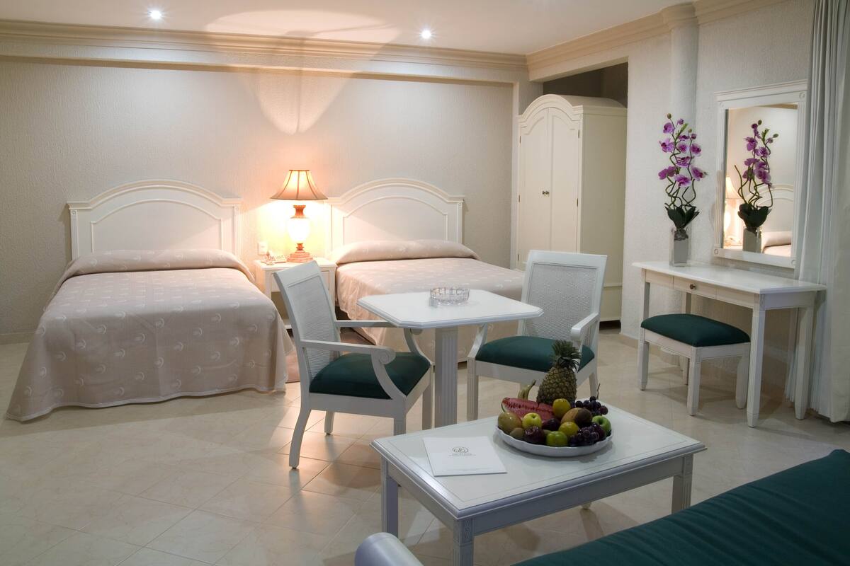 A hotel room with two beds, a small table with a fruit bowl, a desk with a chair, a lamp, and a mirror.