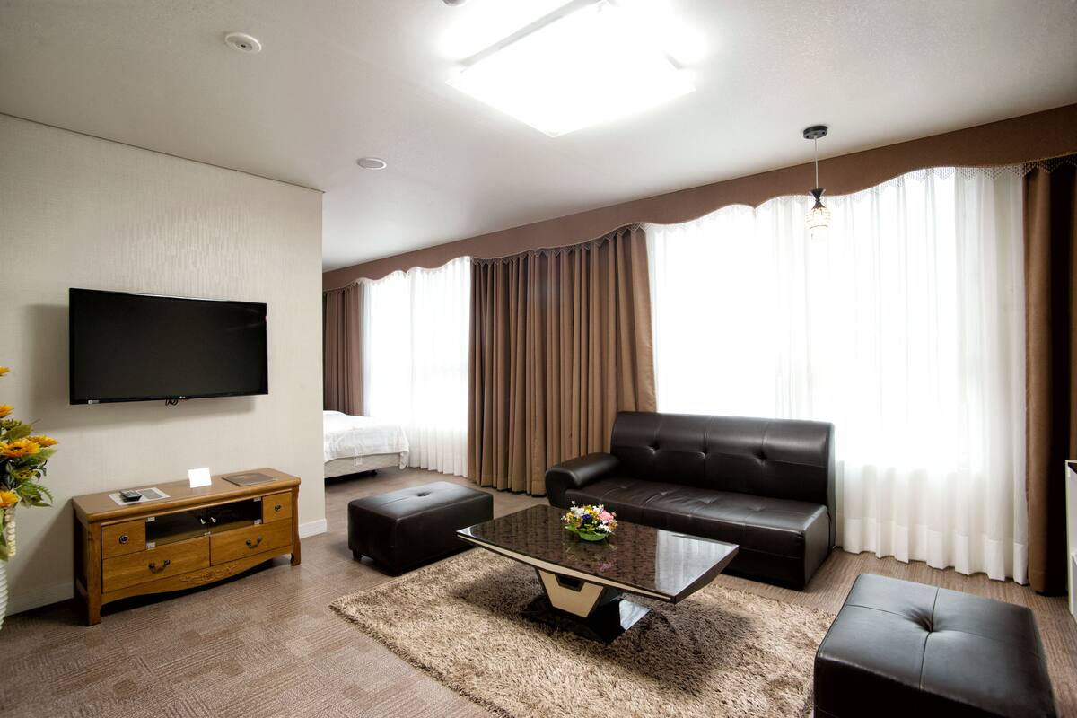 Una habitación de hotel moderna con un televisor de pantalla plana montado en la pared, un sofá de cuero, una mesa de centro de vidrio y una mesita de noche de madera.
