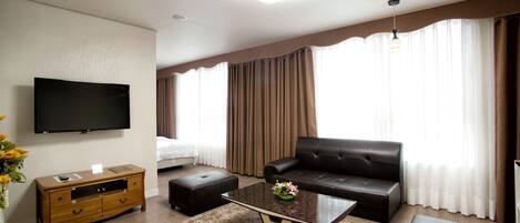 A modern hotel room with a flat-screen TV mounted on the wall, a leather sofa, a glass coffee table, and a wooden side table.