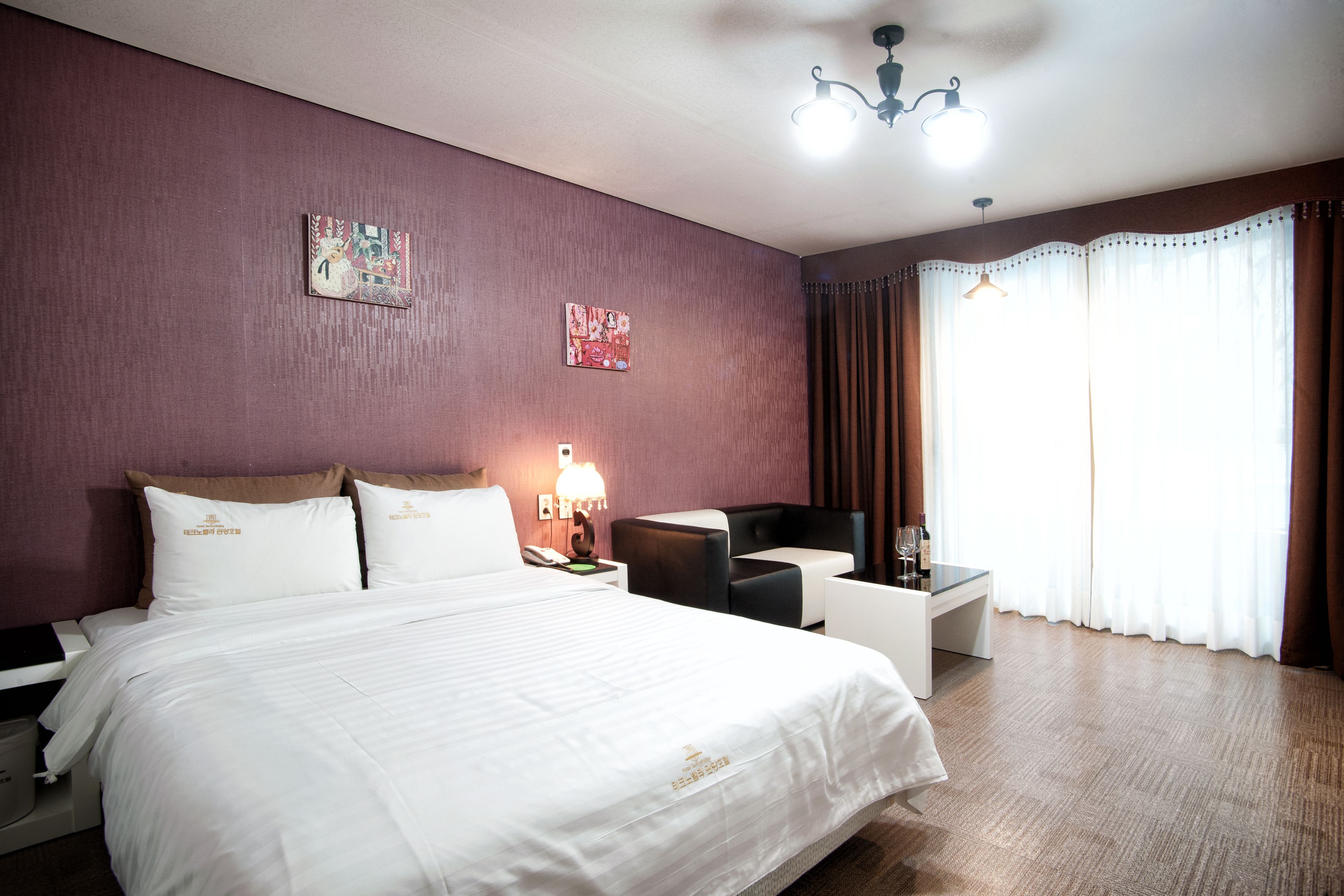 A hotel room with a large bed, a desk, a chair, and a lamp. There are two paintings on the wall and a window with curtains.