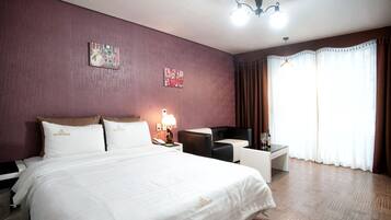 A hotel room with a large bed, a desk, a chair, and a lamp. There are two paintings on the wall and a window with curtains.