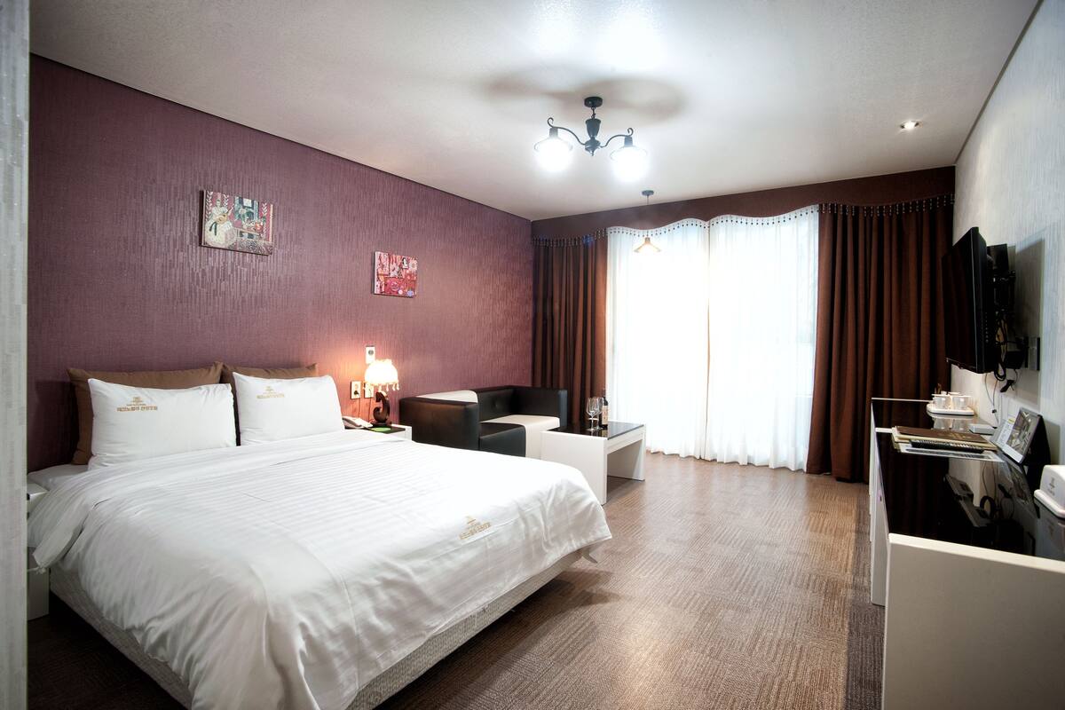 Habitación de hotel con una cama grande, un escritorio, una silla, un televisor y una ventana con cortinas.
