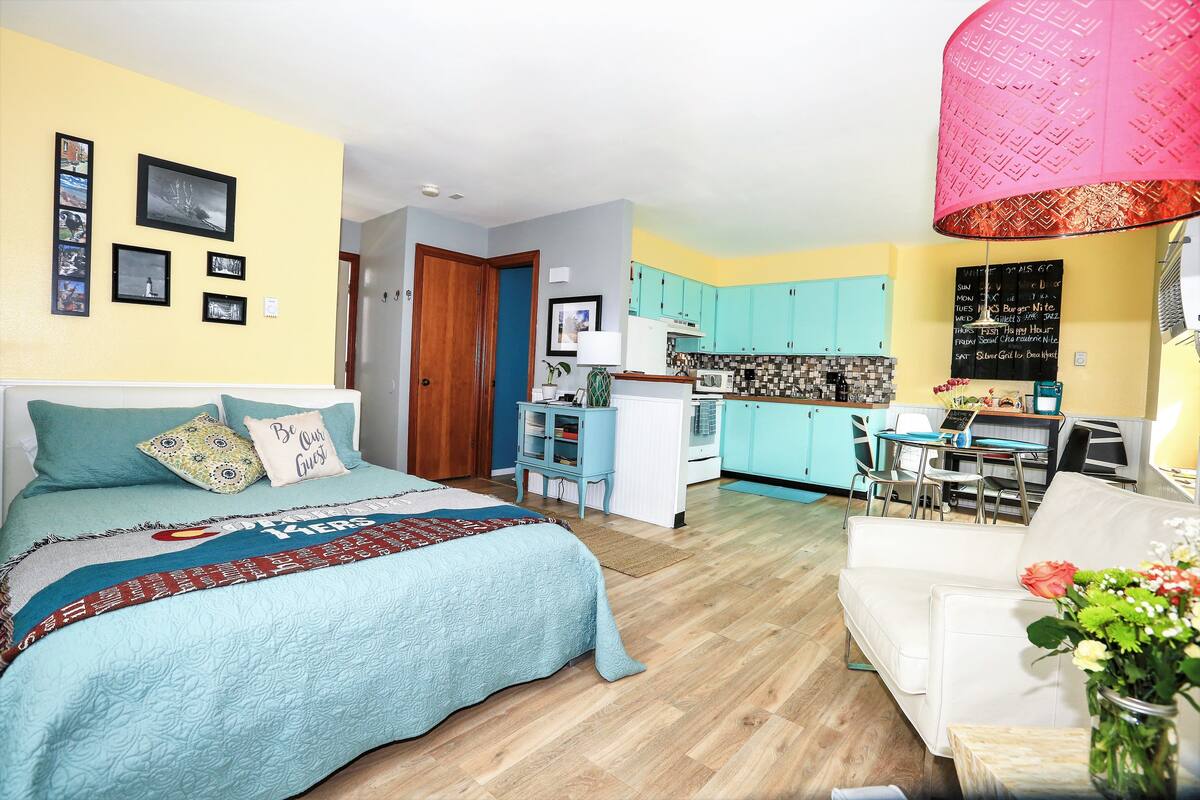 Quarto com cama, mesa de cabeceira e criado-mudo. Há uma janela com cortinas e vista para o exterior.