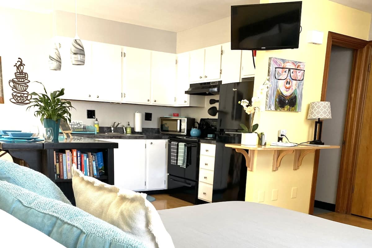 Quarto com cama, TV fixada na parede e área de cozinha ao fundo.