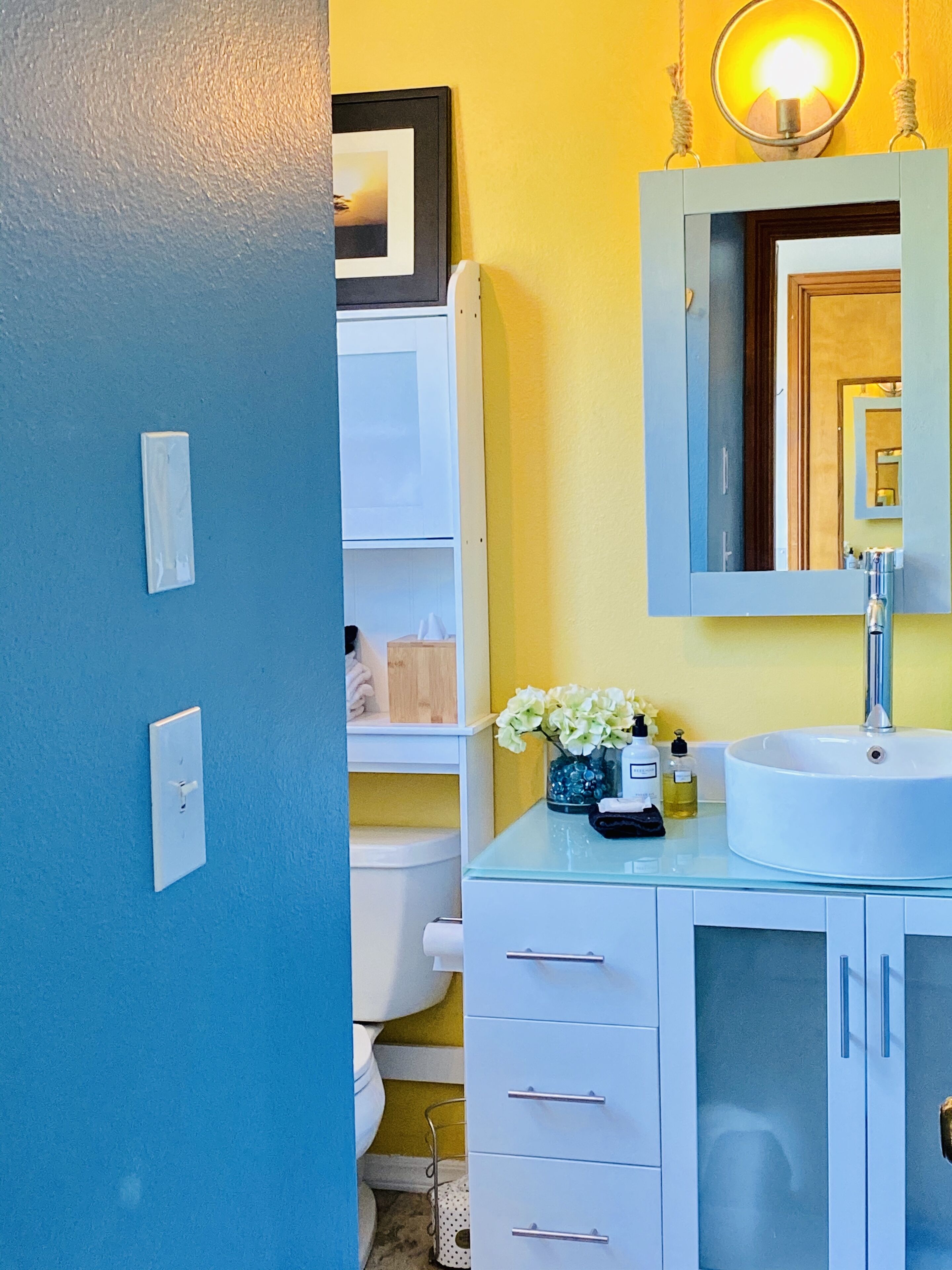 A bathroom with a white sink, a mirror, and a toilet. There is a light fixture above the mirror and a towel rack on the wall.