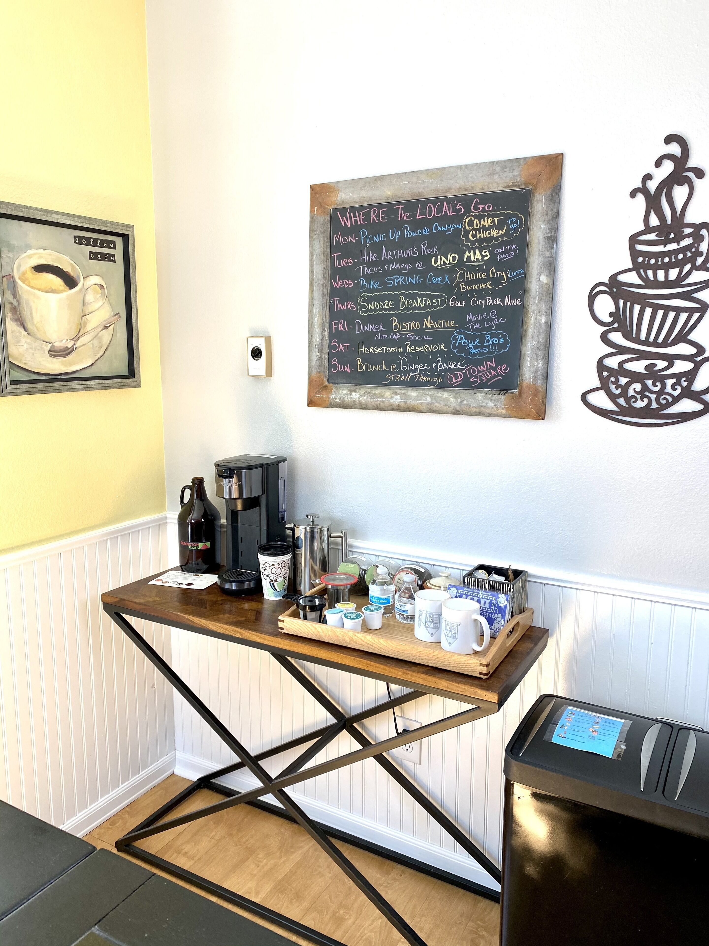 A coffee station with a chalkboard menu listing daily specials, a coffee machine, and various mugs and containers on a wooden table.