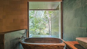 A stone bathtub in a bathroom with a view of a lush garden. - BeingSattvaa Luxury Ubud (Ubud)